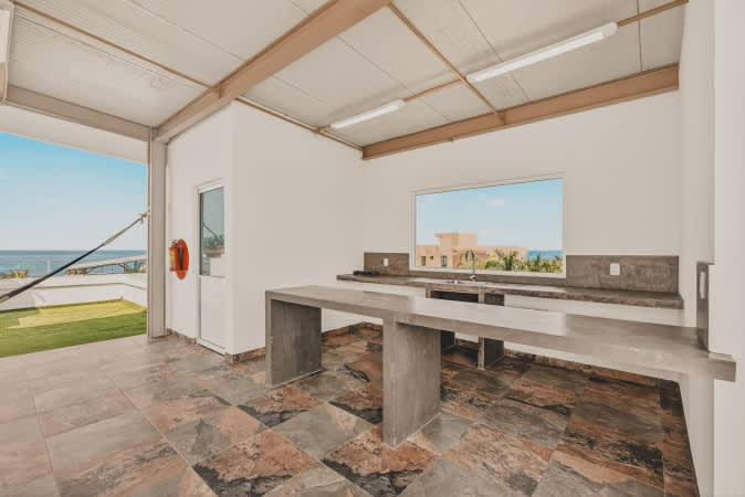 Outdoor kitchen space with concrete counters and a bar, featuring patterned floor tiles and an open side providing a clear view towards the ocean and sky.