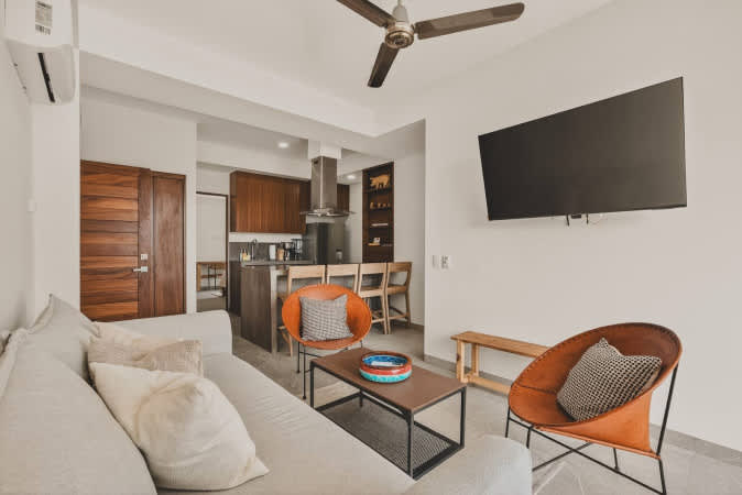 Spacious open-concept living room and kitchen area, featuring a light grey sofa, two orange accent chairs, and a coffee table, with the modern kitchen island and wooden cabinetry visible in the background.