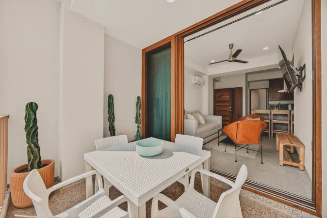 Balcony with a white wicker dining table and chairs, featuring a light blue bowl centerpiece and tall potted cacti. Large sliding glass doors connect to the interior living space.