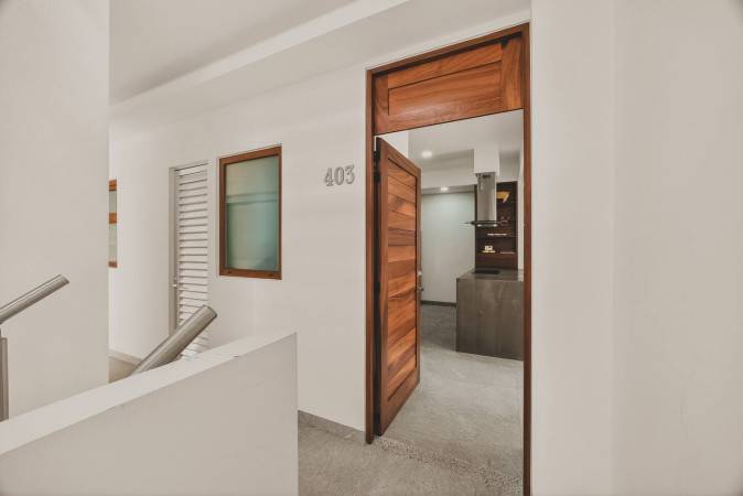 Apartment entrance hallway with a wooden door leading into the unit, marked with '403'. White walls and a light gray floor are visible, with a window and stairs in the background.