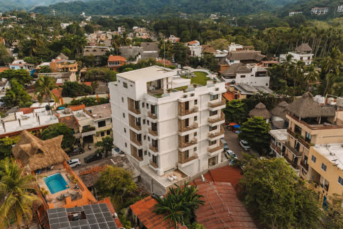 Aerial view of a modern, multi-story white apartment building with balconies, situated within a densely packed town surrounded by lush green hills and other residential structures.