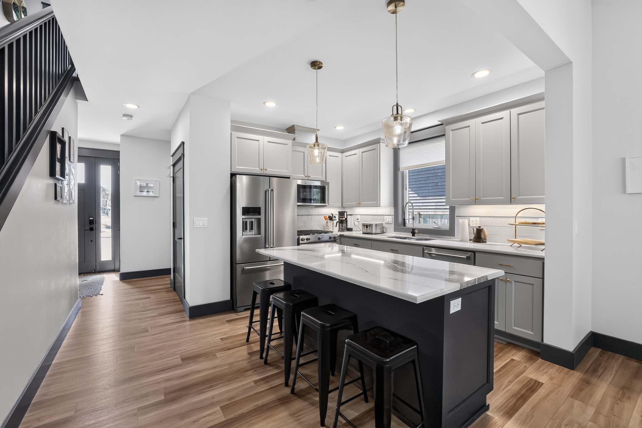 Modern kitchen with light gray shaker cabinets, white subway tile backsplash, and stainless steel appliances, centered around a dark gray island with a white marble countertop and black bar stools. Hardwood floors extend into the entryway.