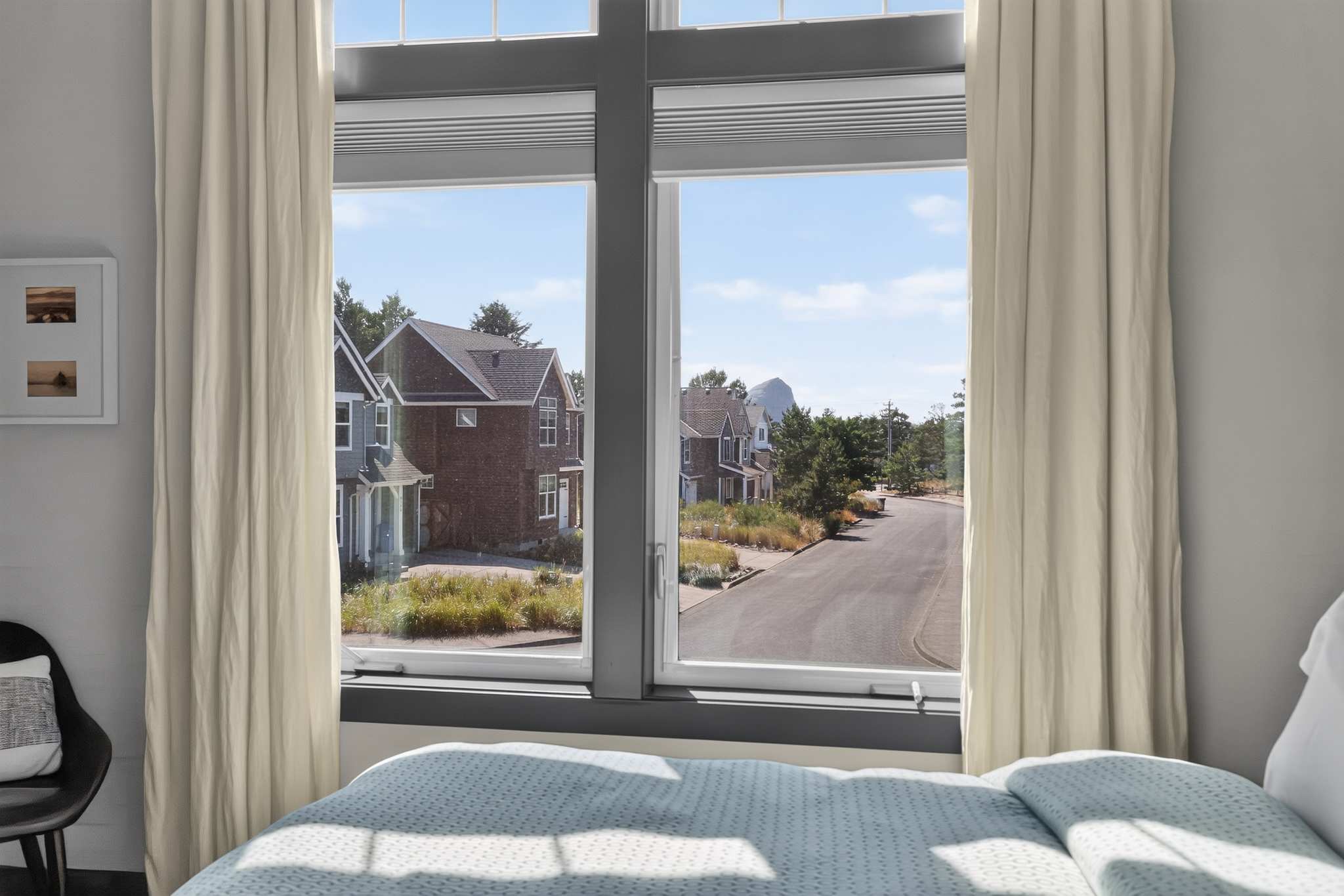 View from a bedroom window with dark gray trim, showcasing a street scene with neighboring houses and lush greenery under a clear sky. White curtains frame the window, offering a glimpse of the surrounding neighborhood.