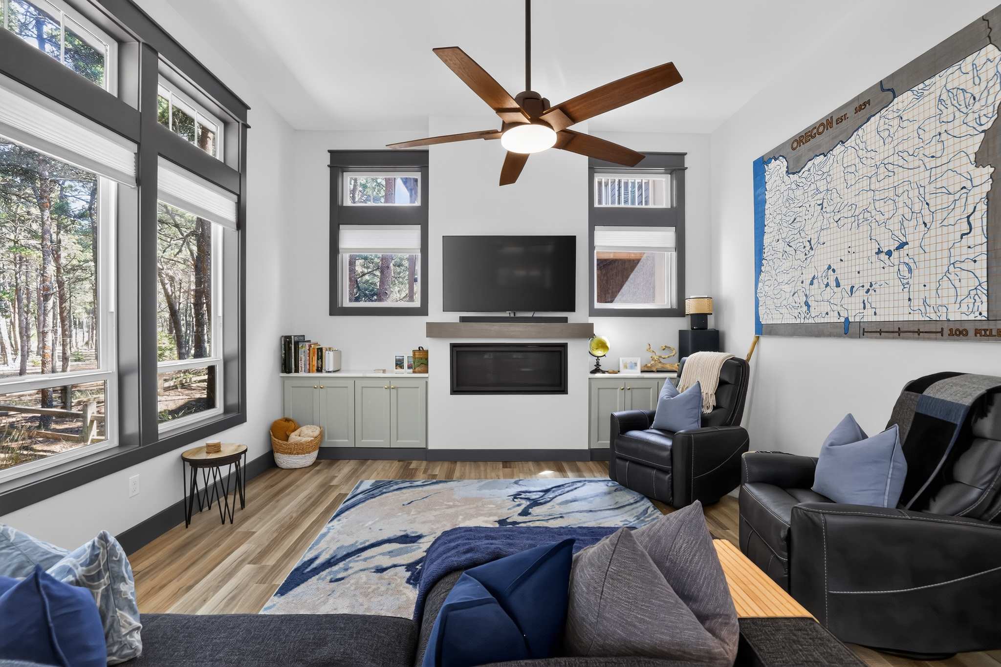 Spacious living room with light walls, dark trim around large windows, a modern fireplace with a mounted TV above, and built-in light green cabinets. The room features two black recliners, a large area rug with blue and white patterns, and a wooden ceiling fan.