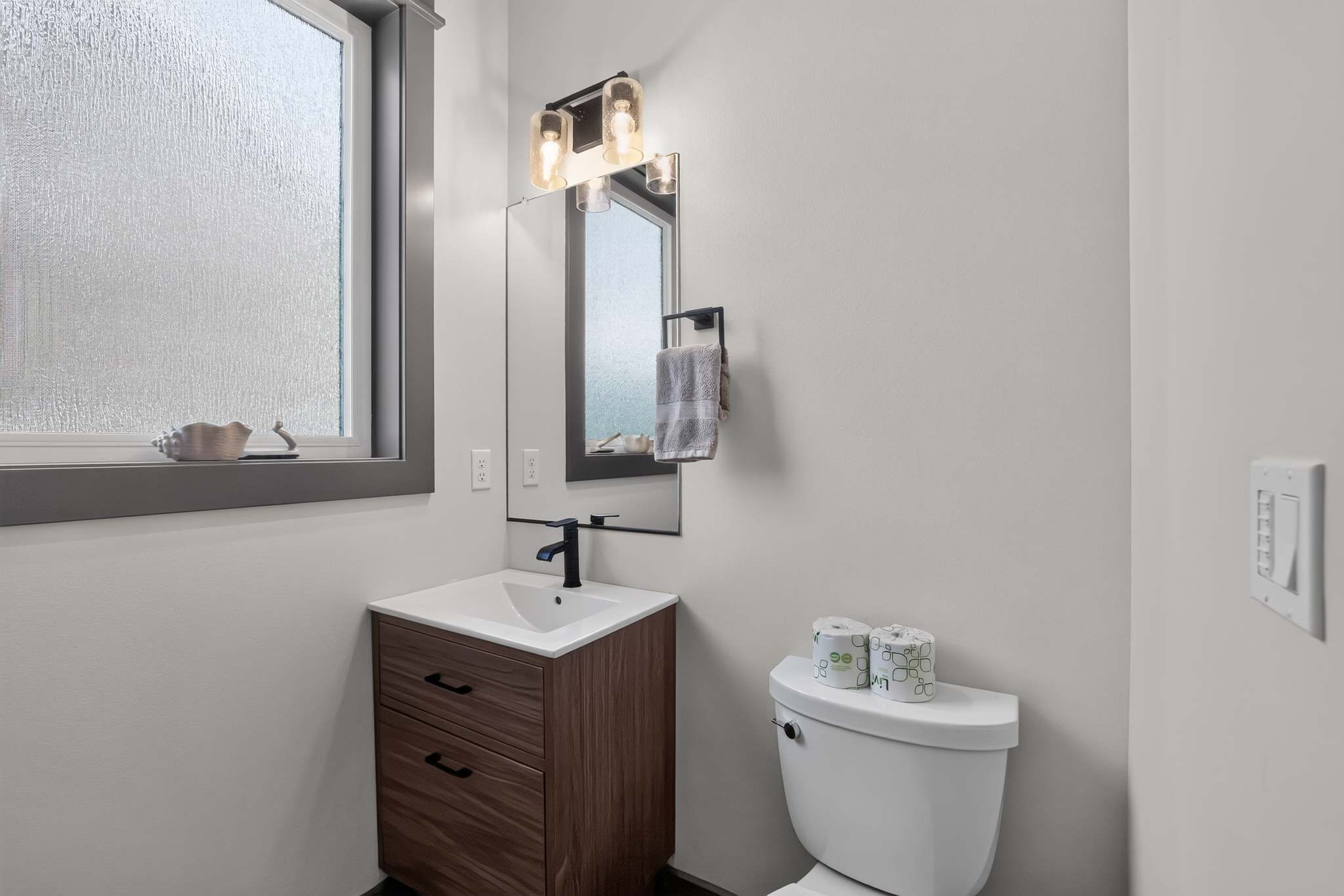 Compact modern half-bathroom featuring a dark wood vanity with a white sink and black faucet, a rectangular mirror, and a toilet. A frosted window provides natural light.