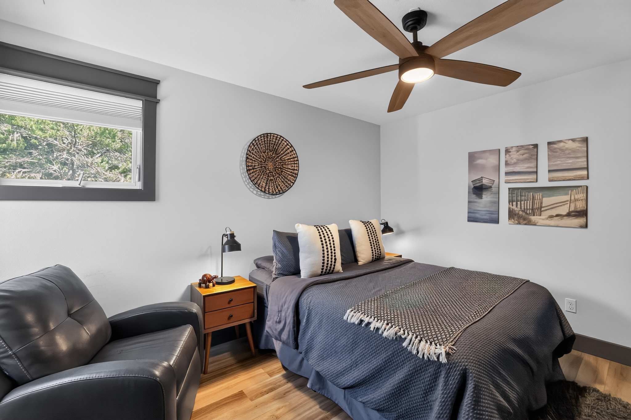 Cozy bedroom with light gray walls, a dark blue bedspread, two nightstands with black lamps, and a large wooden ceiling fan. Wall art includes a woven circular piece and a series of four coastal-themed paintings above the bed.