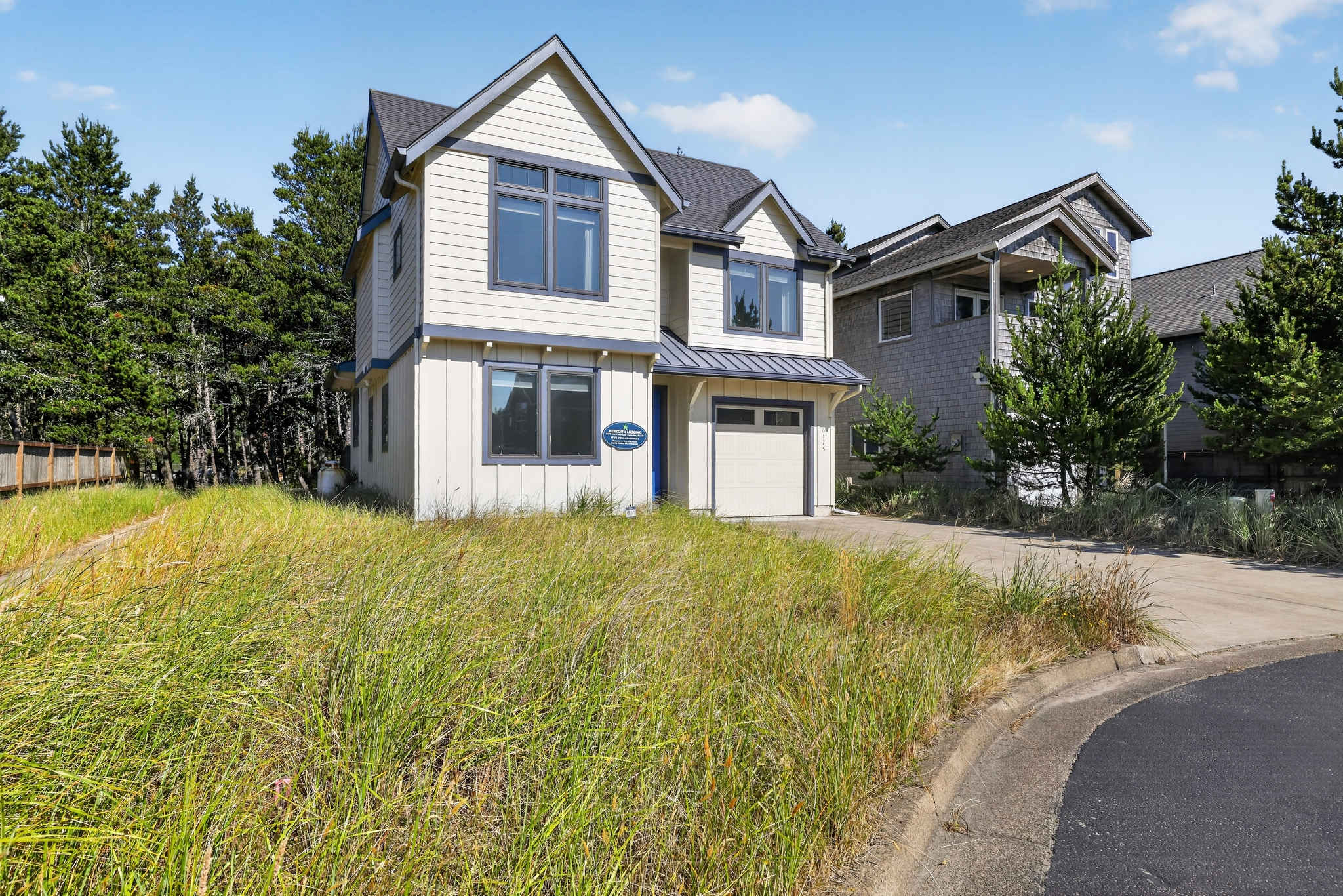 Two-story coastal house with light yellow siding and dark gray trim, featuring a small garage, a front door with a small porch, and a paved driveway. The property is surrounded by tall wild grass and trees, with a curved paved road in the foreground.