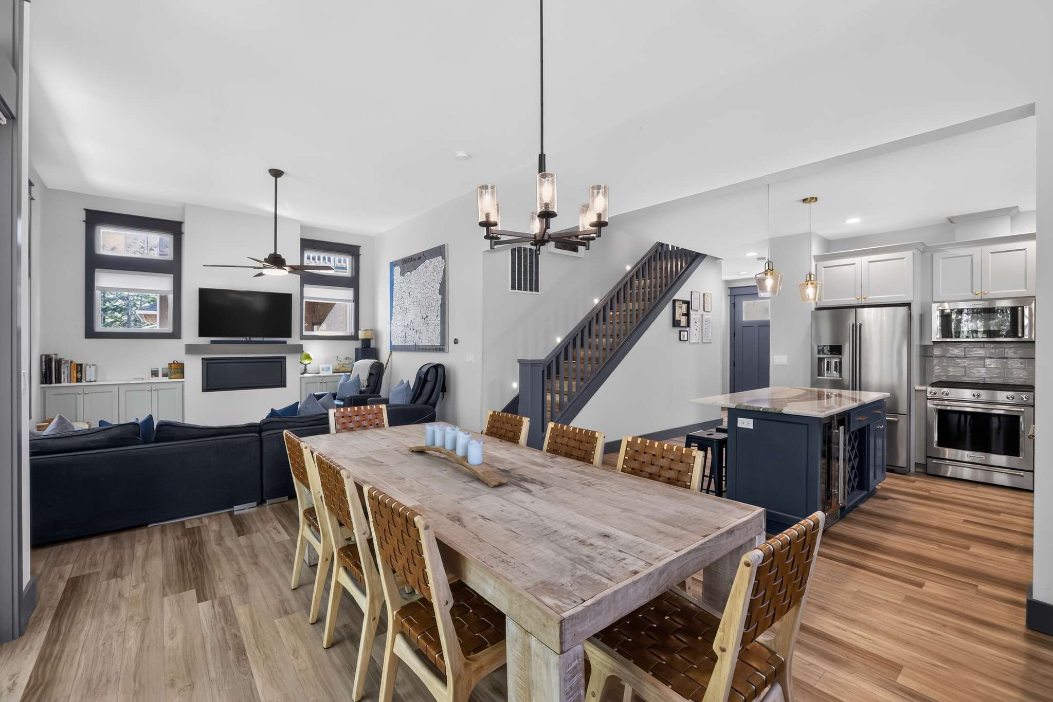 Open-concept living and dining area featuring a large rustic wooden dining table with eight leather-seated chairs, leading into a cozy living room with a dark blue sectional sofa and a wall-mounted TV above a fireplace. A dark staircase is visible in the background.