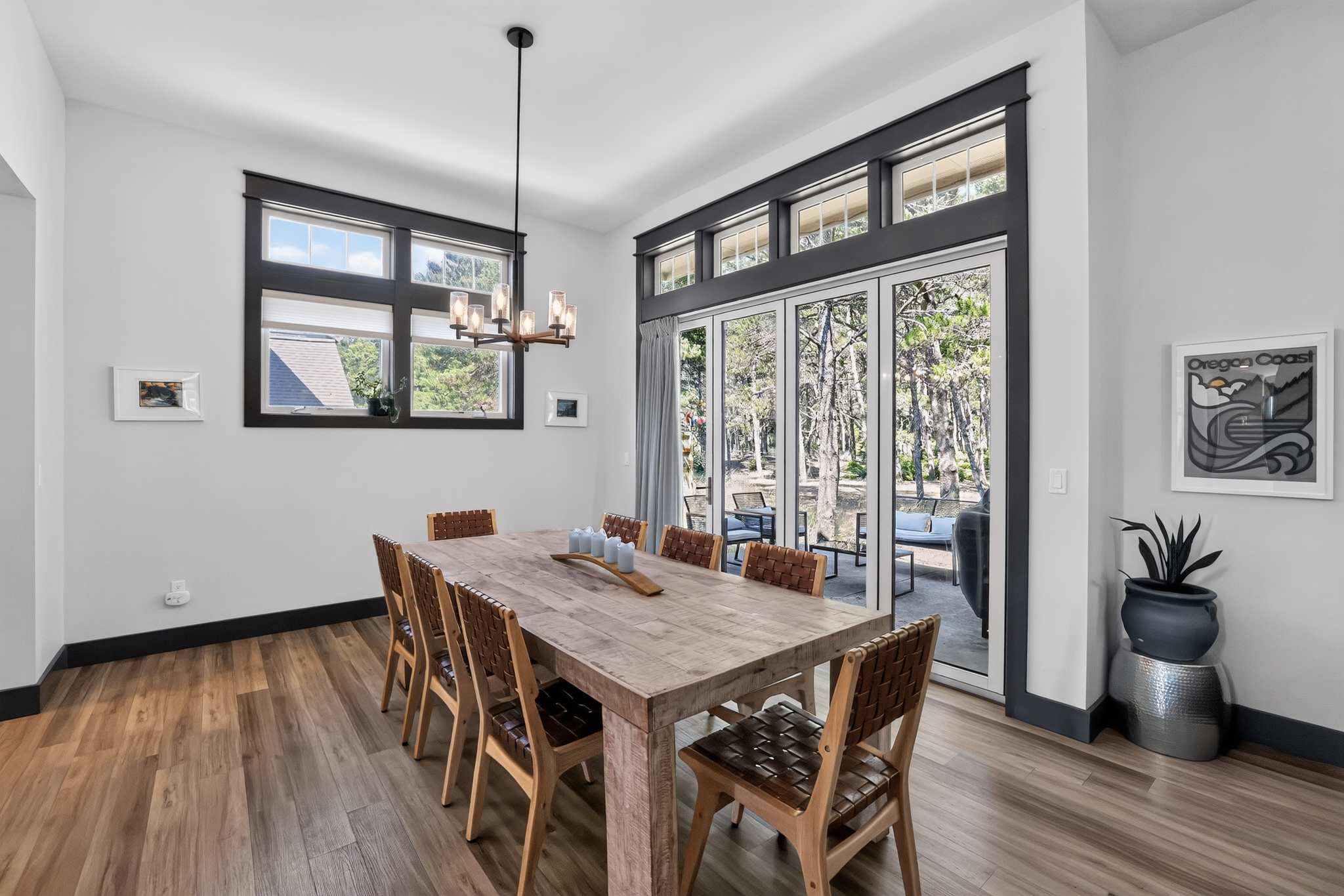 Elegant dining room featuring a large rustic wooden table with eight woven leather chairs, illuminated by a modern chandelier. Large windows and glass doors provide ample natural light and views of the surrounding trees, with dark trim accentuating the openings.