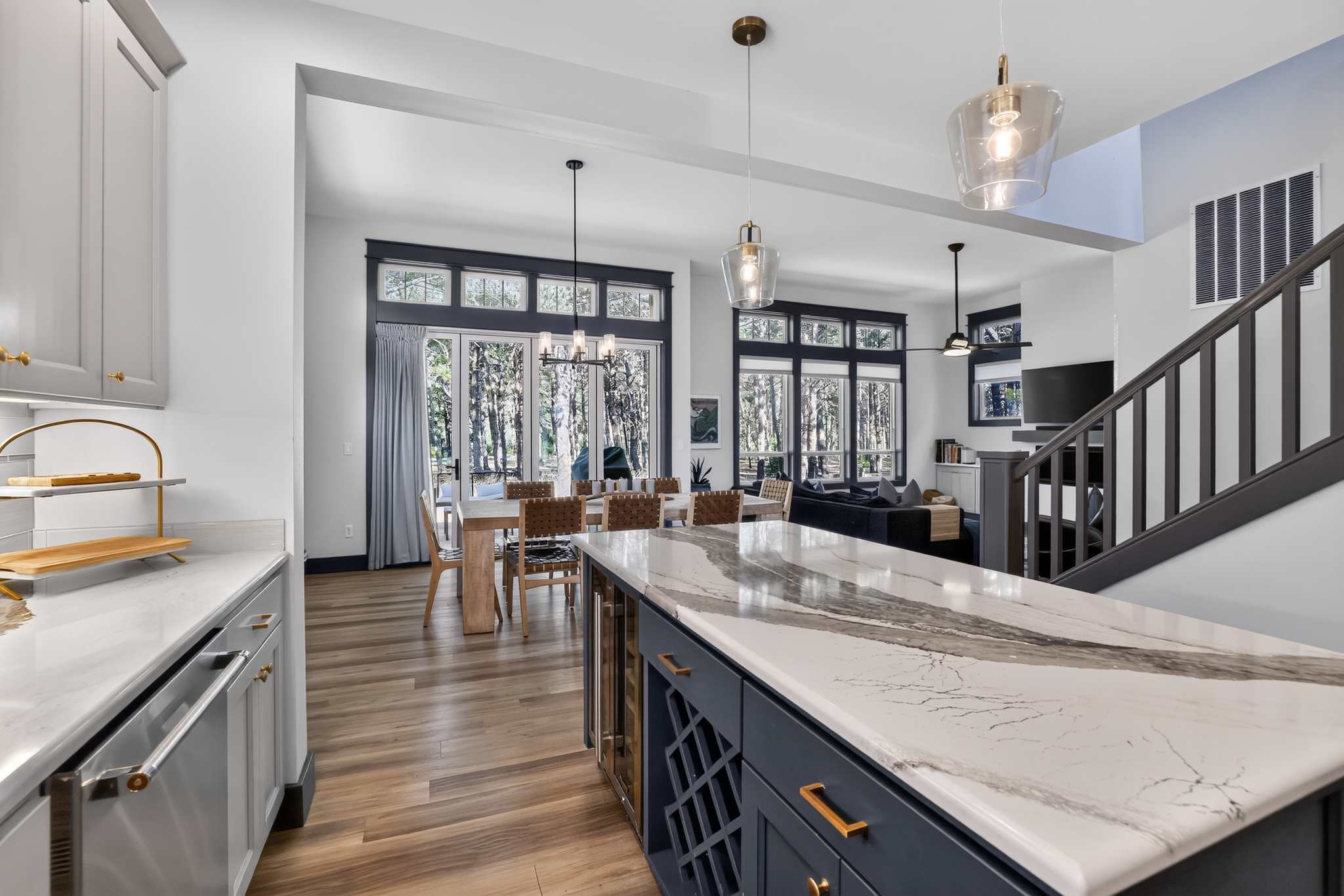 Bright open-concept dining and living area with large windows overlooking a wooded area, featuring a light wooden dining table with woven chairs and a dark blue sectional sofa. A kitchen island with a wine rack is visible in the foreground.