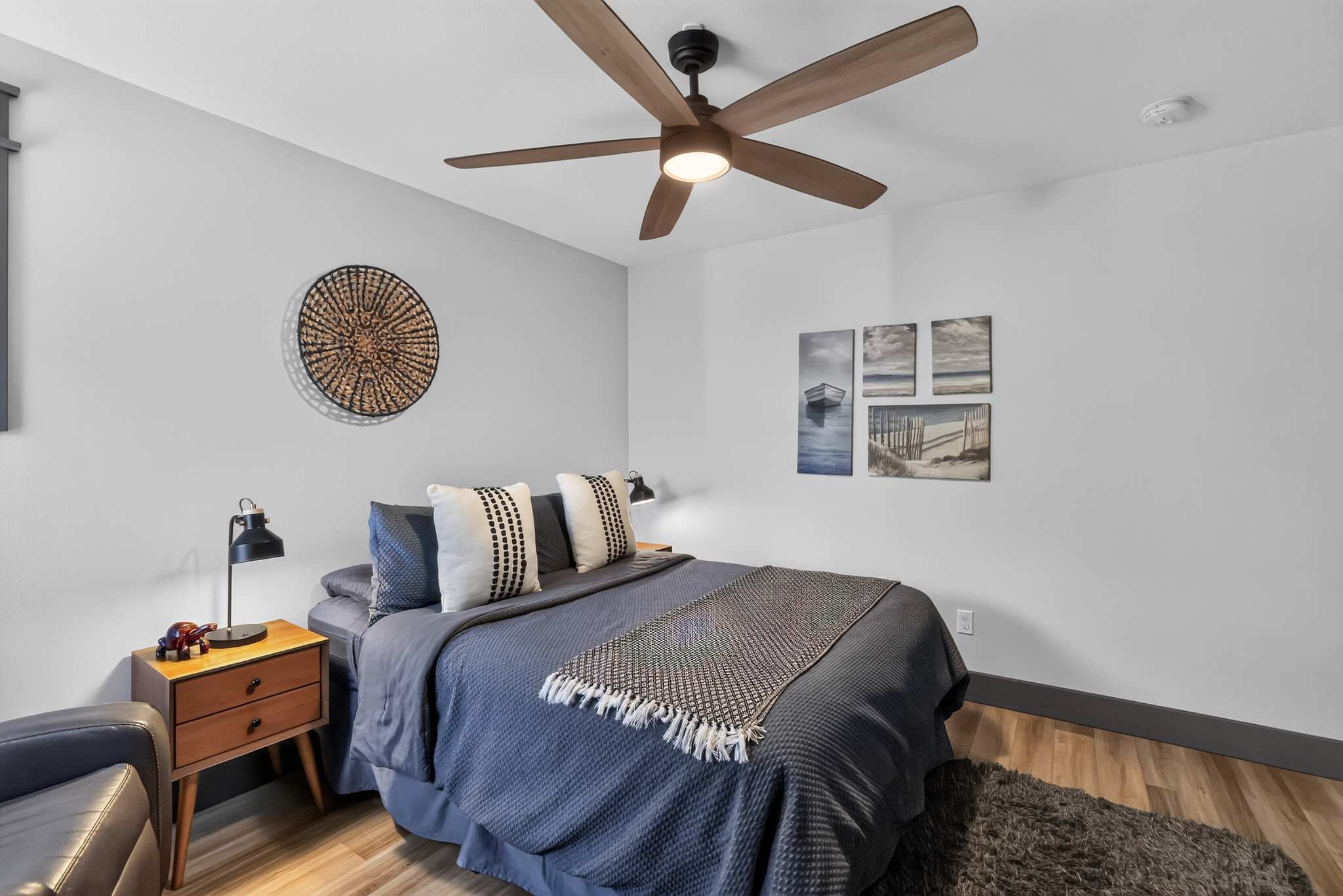 Cozy bedroom with light gray walls, a dark blue bedspread, two nightstands with black lamps, and a large wooden ceiling fan. Wall art includes a woven circular piece and a series of four coastal-themed paintings above the bed.