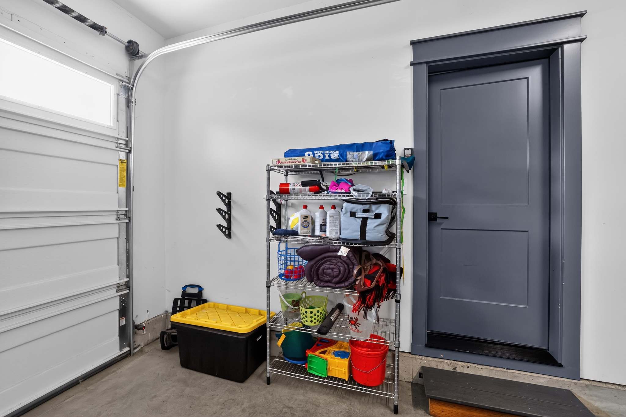Organized garage space with a metal shelving unit holding various household items and cleaning supplies, a yellow storage bin, and a dark gray interior door. The garage door is partially visible on the left.