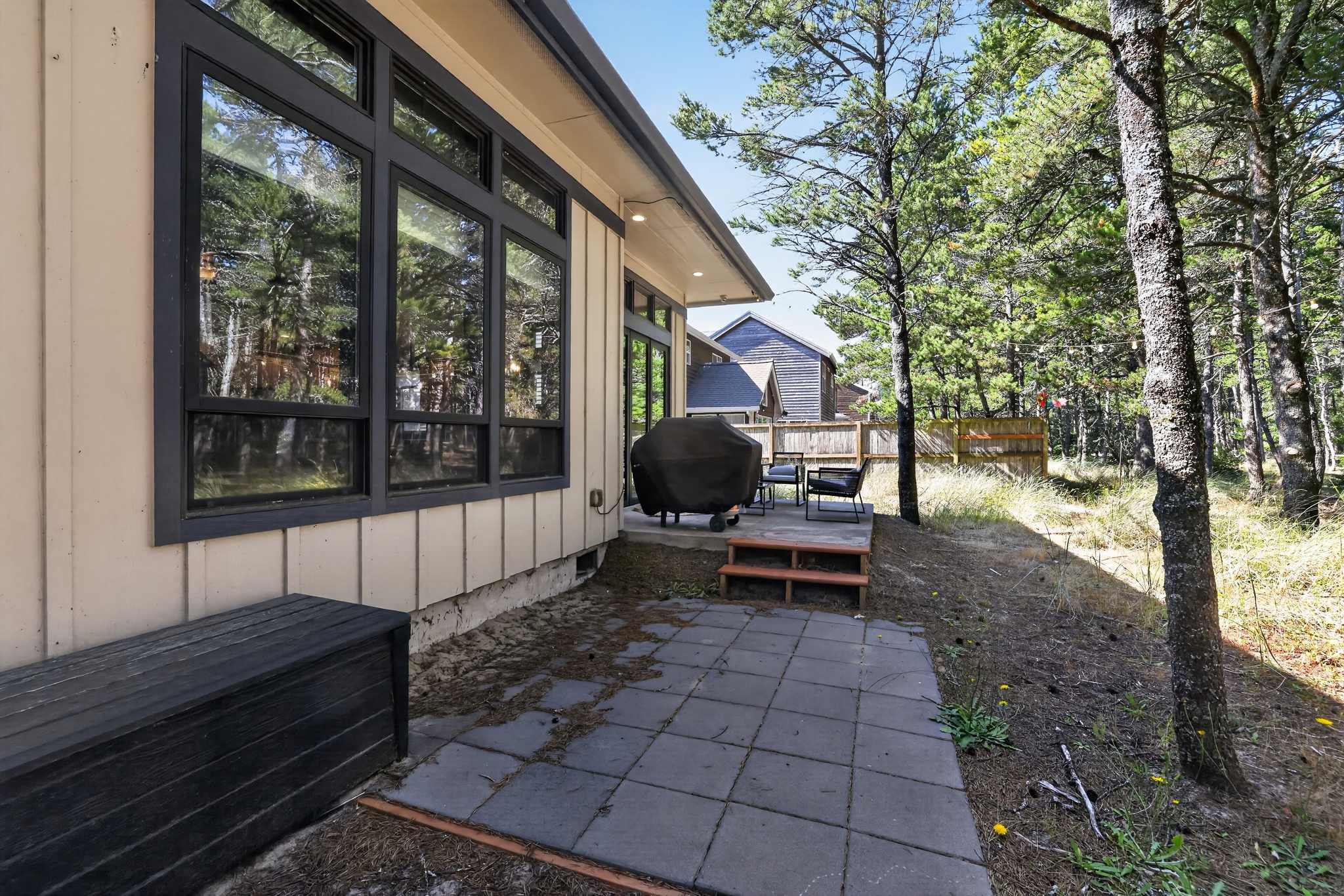 Back exterior of the house featuring a paved patio area with a grill and outdoor seating, surrounded by natural landscaping and tall pine trees. The house has light-colored siding and large dark-framed windows.