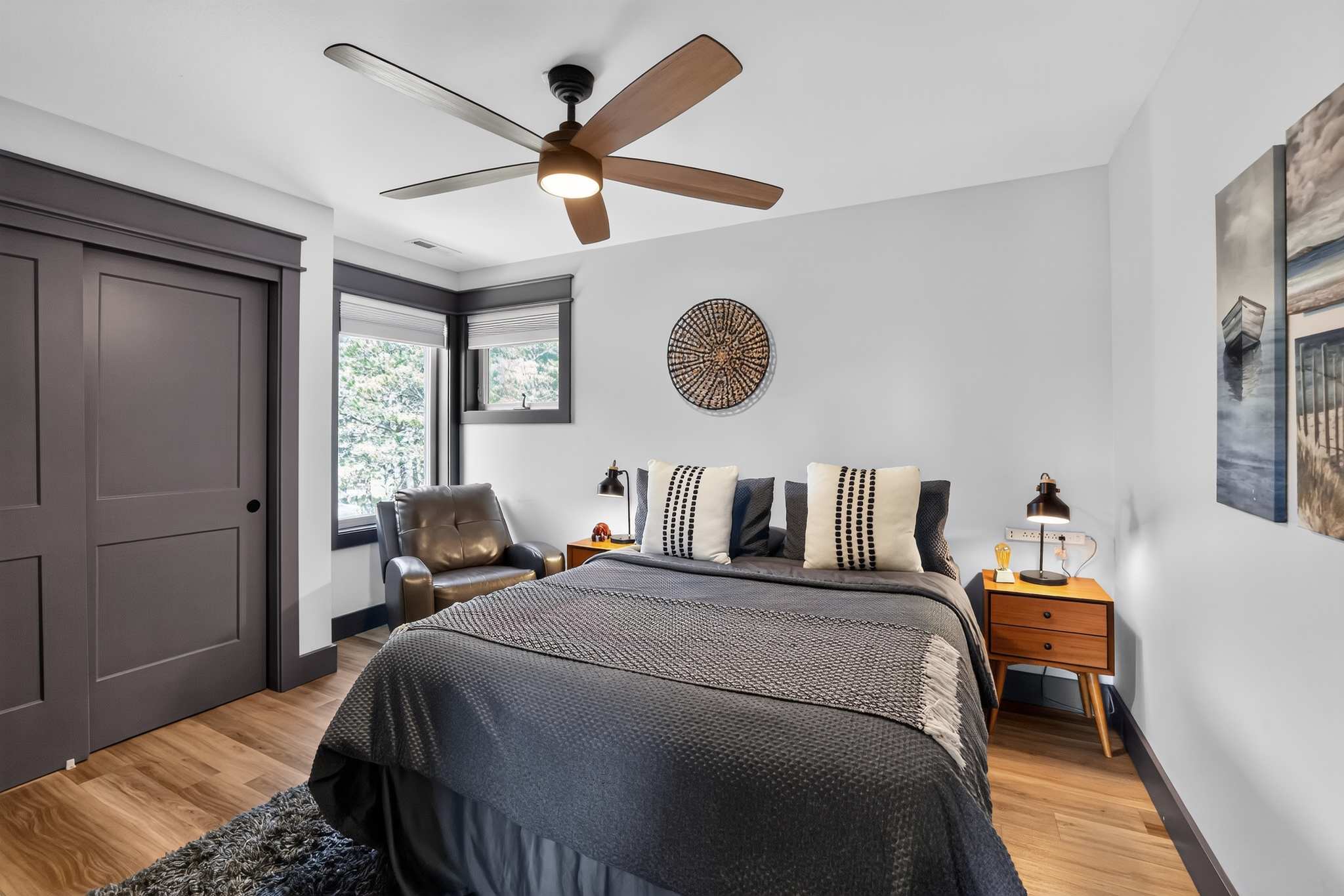 A modern bedroom with light gray walls and dark trim features a queen-sized bed with dark gray bedding and decorative pillows, flanked by wooden nightstands with lamps. A brown leather armchair sits by a window, and a large circular woven art piece hangs above the bed.