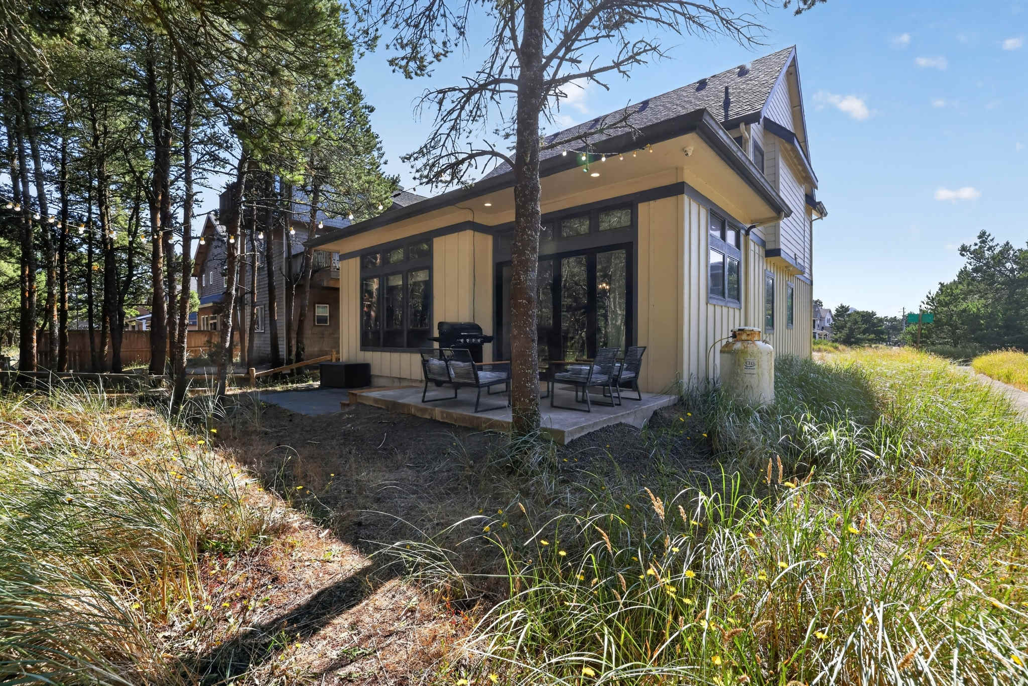 The back of a light yellow house with dark trim, featuring a concrete patio furnished with outdoor seating and a grill. Tall pine trees surround the property, and a large propane tank is visible on the side.