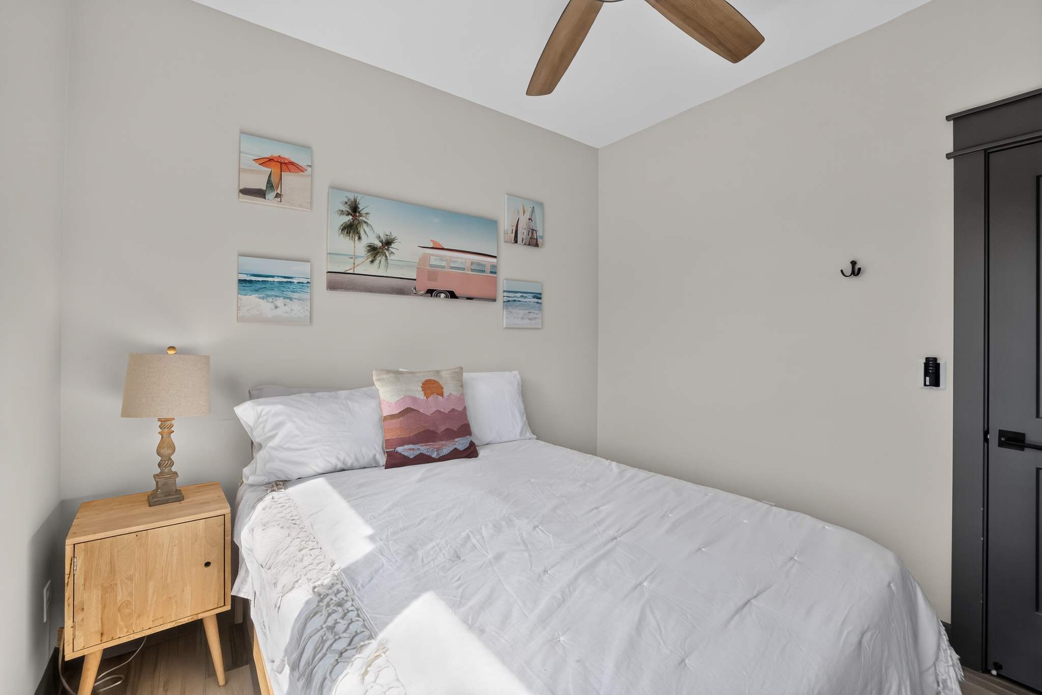 A cozy bedroom with light gray walls features a queen-sized bed with white bedding and a colorful throw pillow, flanked by a light wood nightstand with a lamp. Beach-themed artwork, including a pink vintage van, adorns the wall above the bed.
