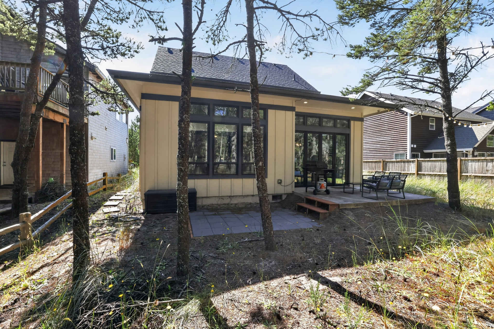 A wider view of the light yellow house's rear, showcasing two distinct patio areas with outdoor furniture and a grill. The property is nestled among tall pine trees, with a small wooden fence visible on the left.