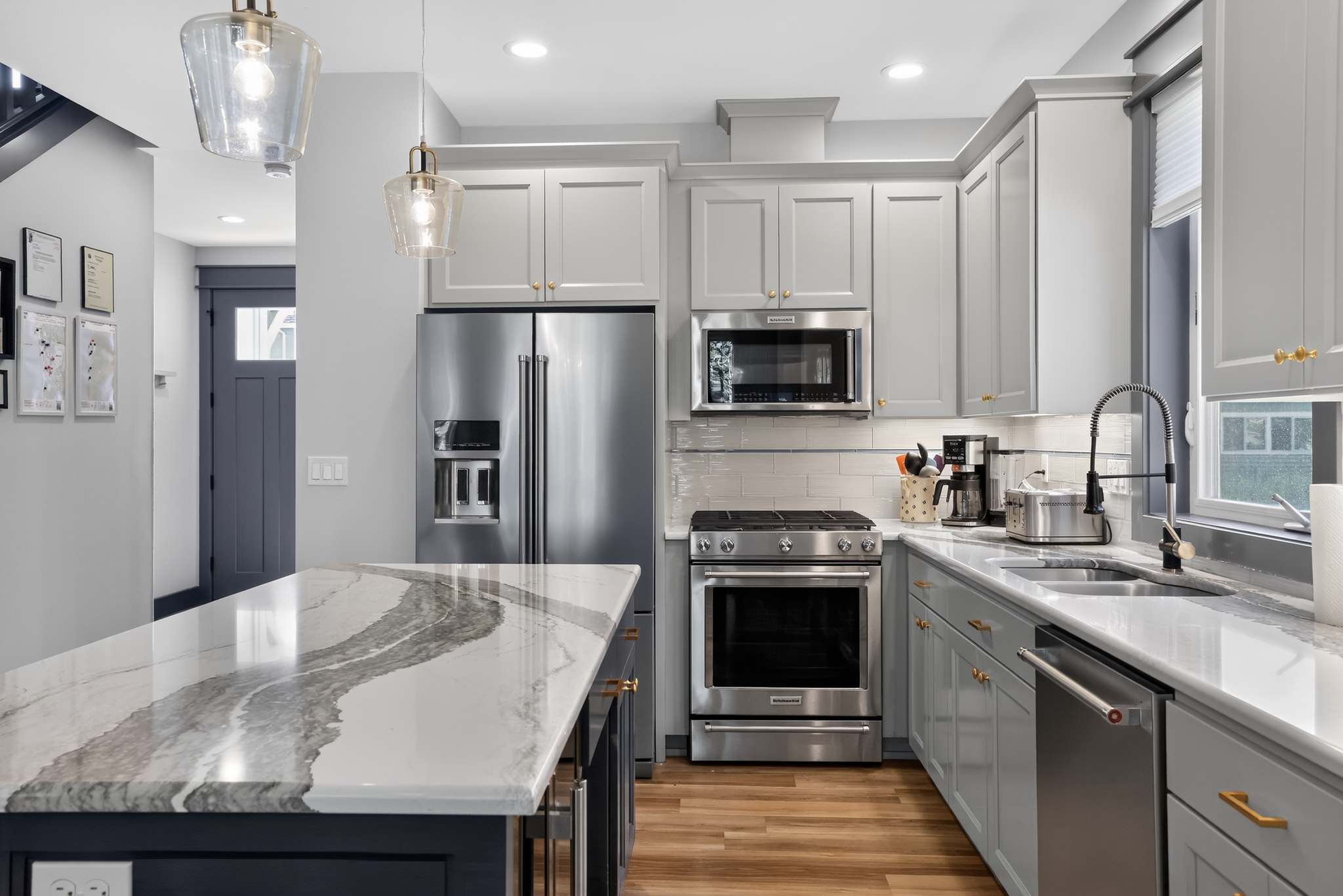 Modern kitchen featuring light gray shaker cabinets with gold hardware, stainless steel appliances including a French door refrigerator, gas range, and microwave, a white subway tile backsplash, and a large island with a dramatic gray and white veined marble countertop.