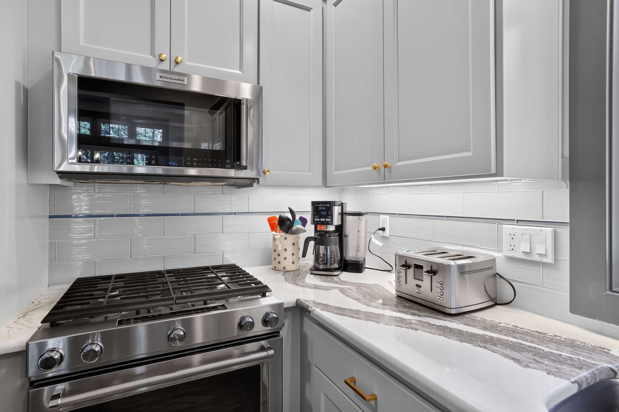 A modern kitchen corner features light gray cabinets with gold hardware, white subway tile backsplash, a stainless steel gas range, and an over-the-range microwave, with a coffee maker and toaster on the quartz countertop.