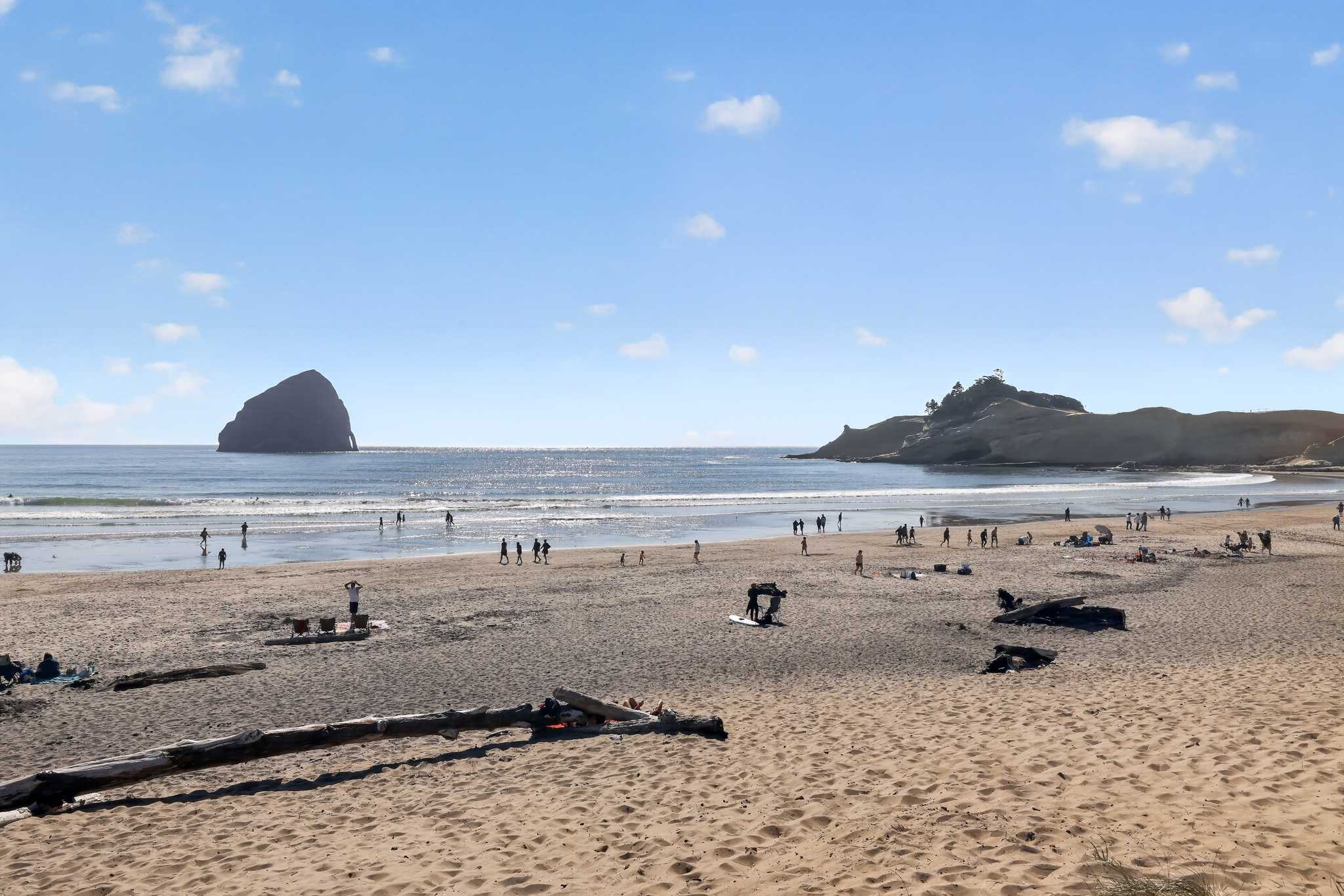Scenic view of a wide sandy beach with the ocean stretching to the horizon, featuring a prominent rock formation in the water and a distant headland. Several people are visible enjoying the beach.