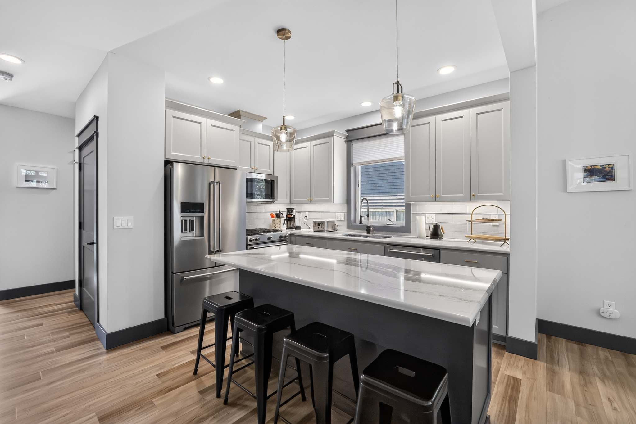 Modern kitchen with light gray shaker cabinets, stainless steel appliances, and white marble countertops. A large island with black bar stools serves as a breakfast bar, illuminated by two pendant lights, and features a subway tile backsplash.