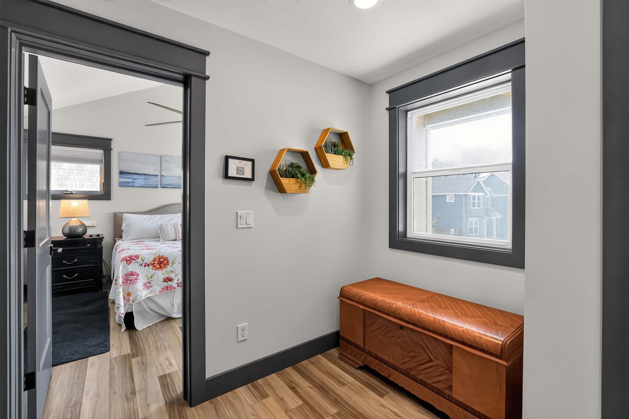 A cozy transitional space features a window with dark trim, a vintage leather-topped wooden chest, and two hexagonal wooden shelves with plants, leading into a bright bedroom through an open doorway.