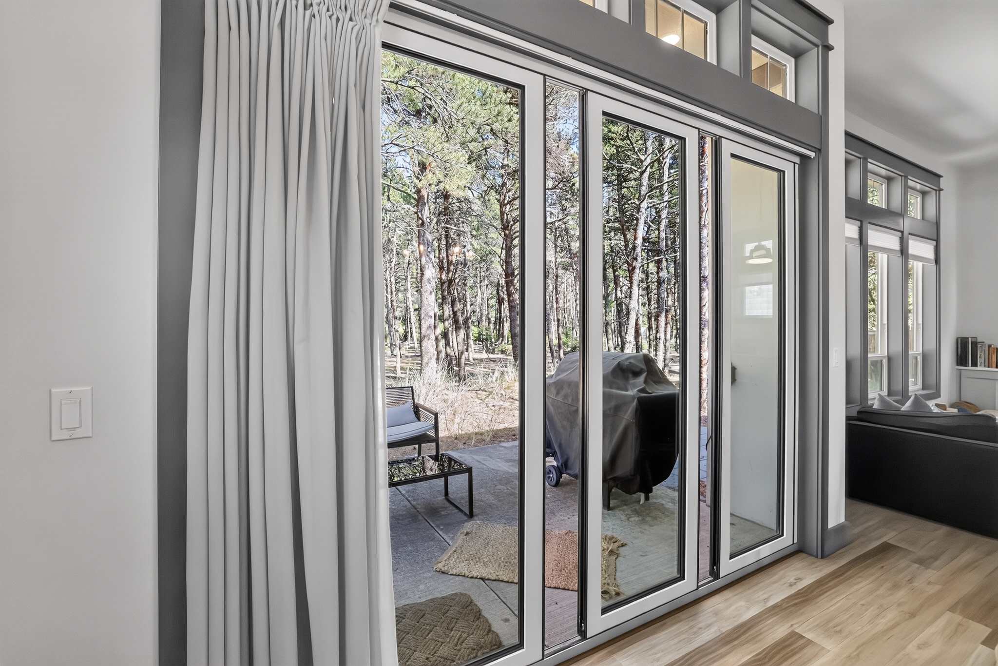 Large multi-panel sliding glass doors open to an outdoor patio area with a grill and seating, surrounded by tall pine trees, bringing natural light and outdoor views into the interior space.
