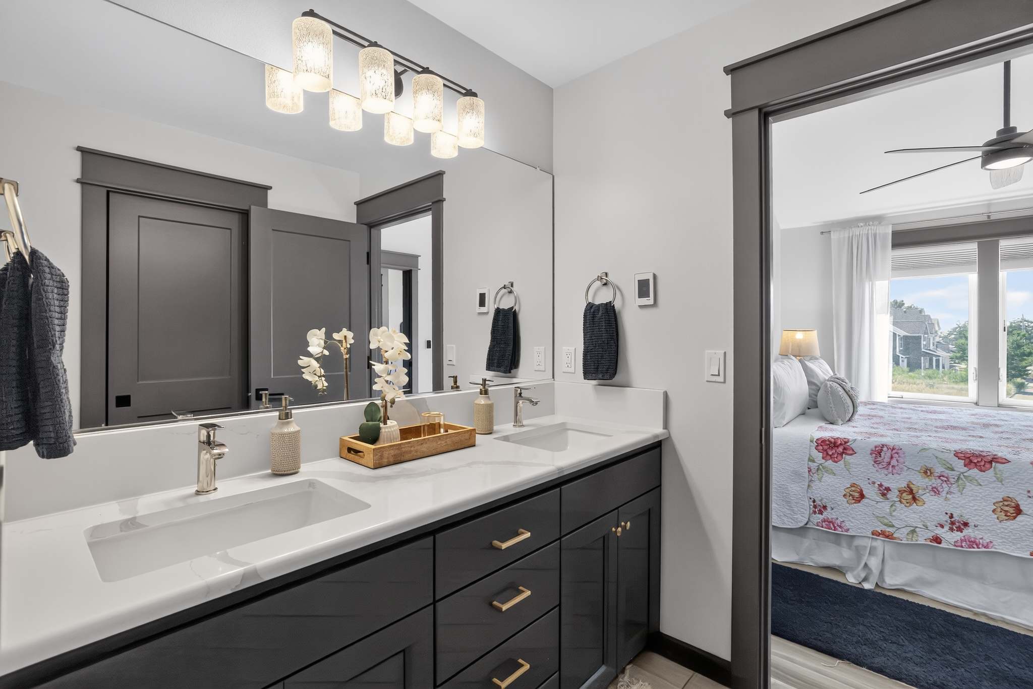 Modern bathroom with a double vanity featuring dark gray cabinets, white marble countertops, and gold hardware, illuminated by a contemporary light fixture above a large mirror. A dark gray barn door leads to an adjacent room, possibly a bedroom.