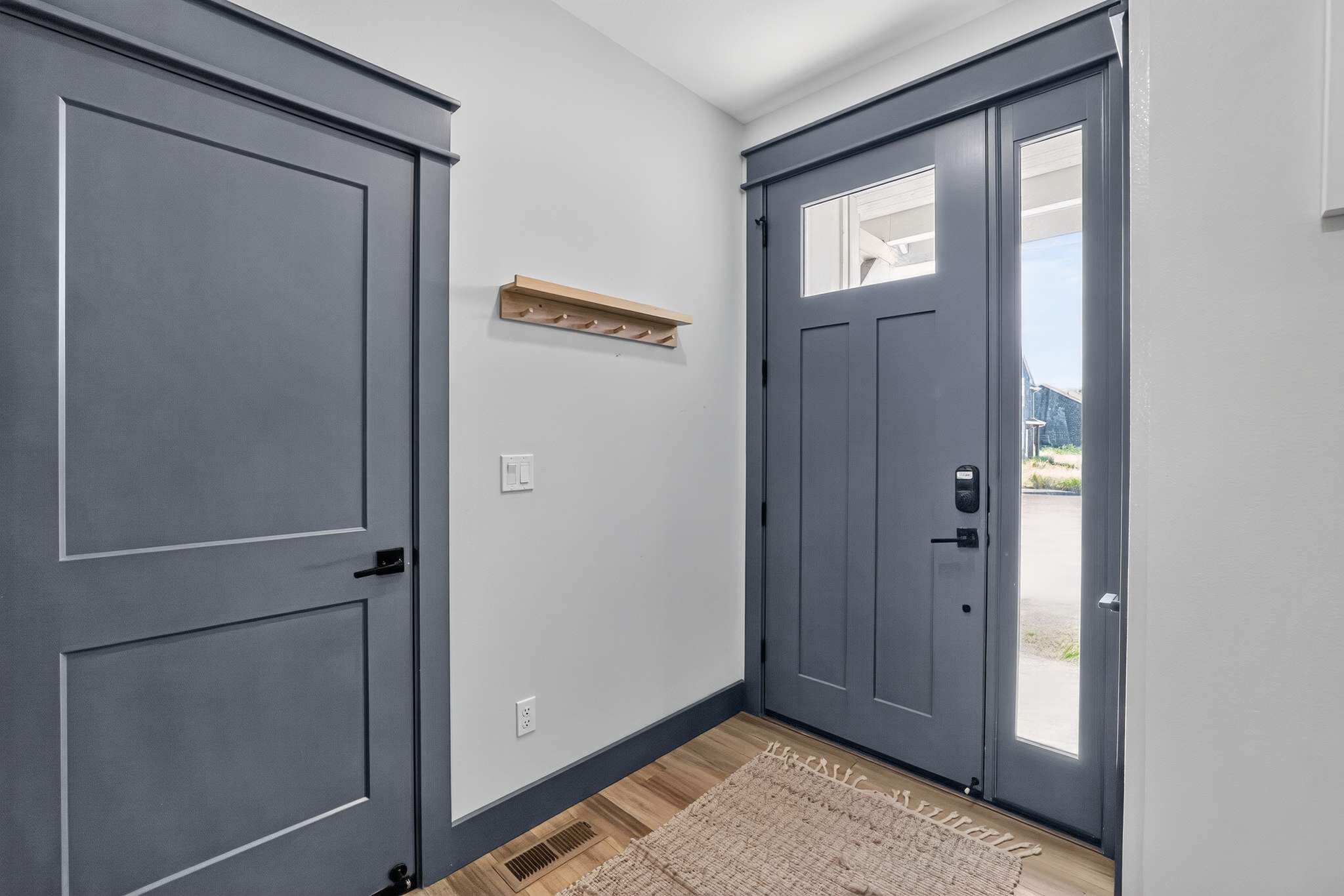Welcoming foyer with light gray walls and dark gray trim, featuring a dark gray front door with glass panels and a matching closet door. A small wooden coat rack is mounted on the wall, and a woven rug rests on the hardwood floor.