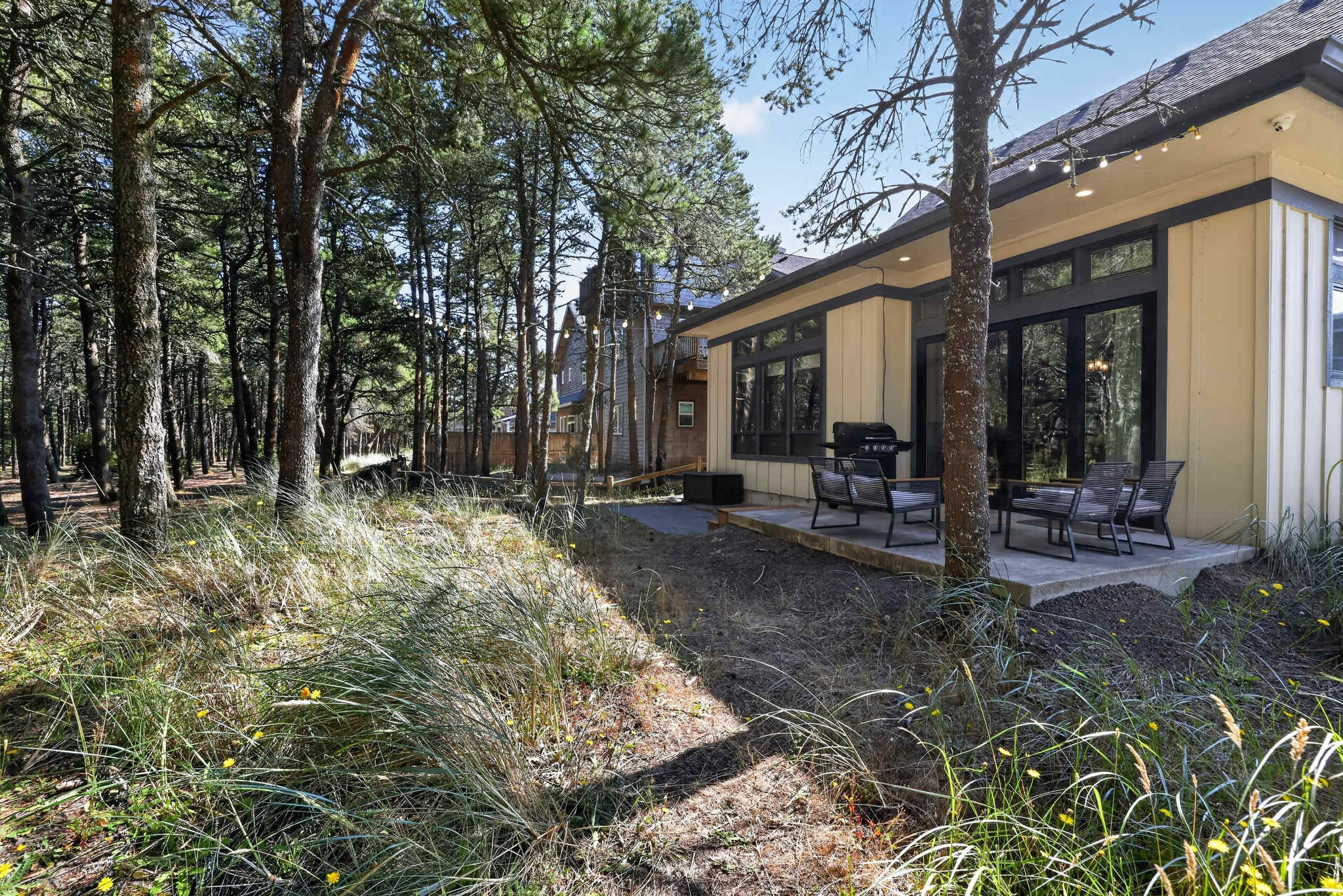 A view of the back of the house, featuring a light yellow exterior with large windows and glass doors, opening onto a concrete patio with outdoor seating. The house is surrounded by tall pine trees and natural vegetation.