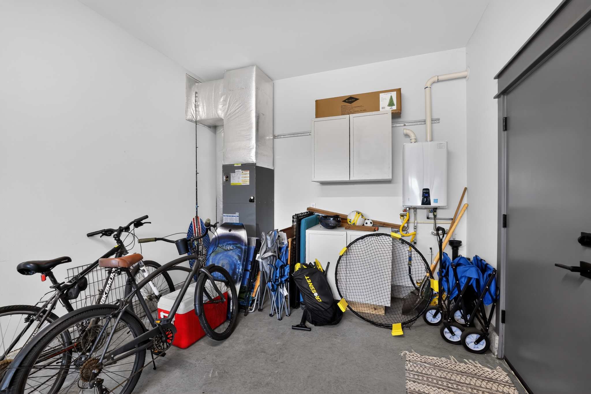 A functional garage space is used for storage, housing bicycles, various outdoor recreational equipment, and utility items, with a water heater and HVAC unit visible against the white walls.