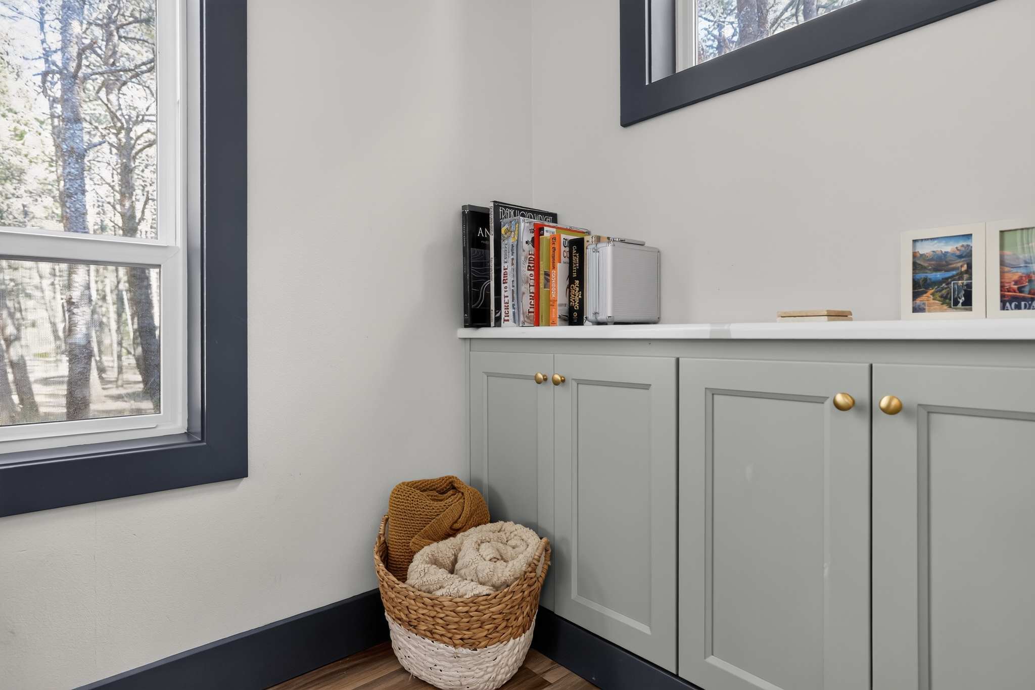 A small nook with light gray walls and dark trim features light gray cabinets with gold pulls, a white countertop, and a woven basket filled with blankets. A window with dark trim looks out onto trees.