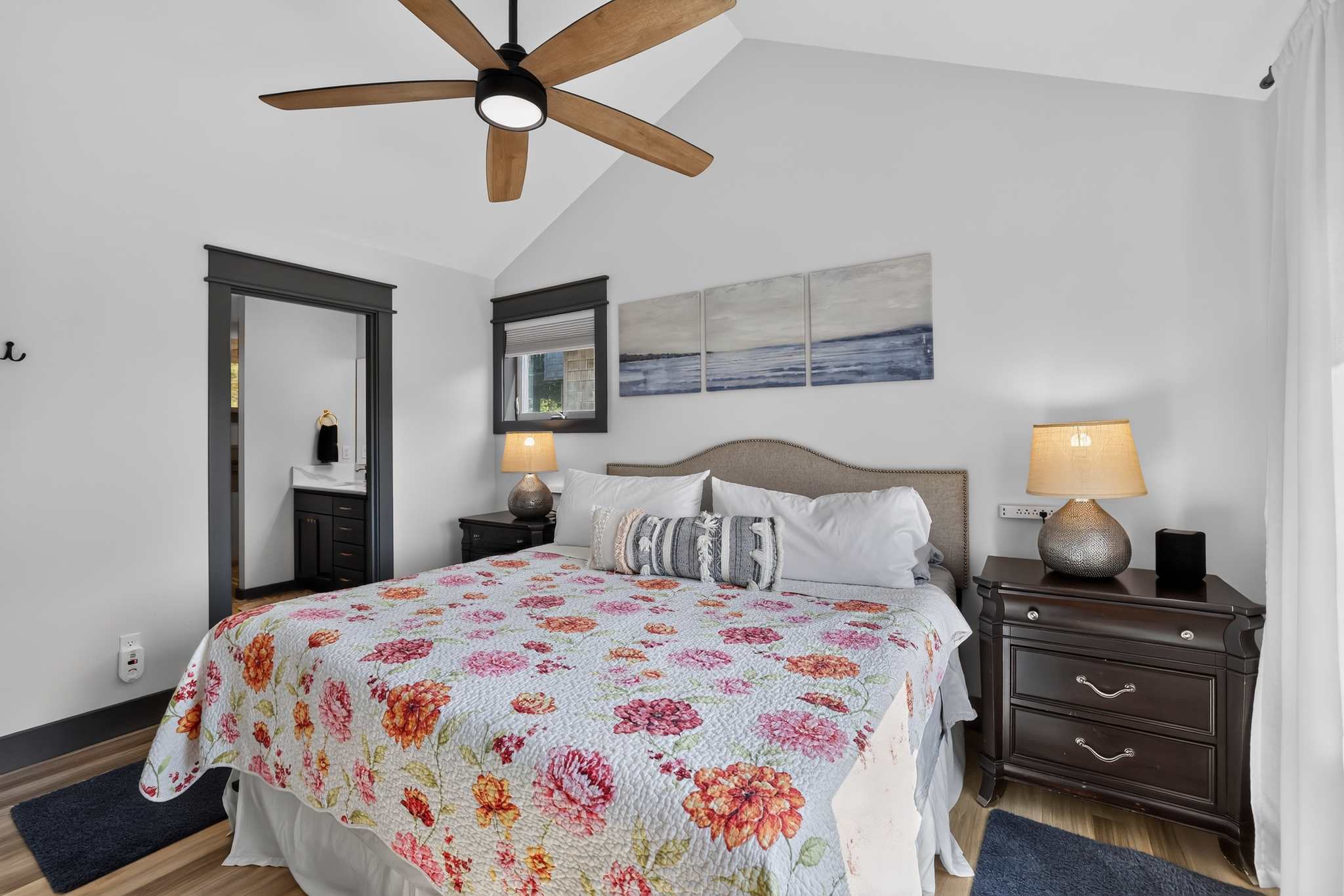 Bright bedroom with a queen-sized bed covered in a vibrant floral quilt, flanked by dark wood nightstands with lamps. The room has vaulted ceilings, a wooden ceiling fan, and a view into an en-suite bathroom.