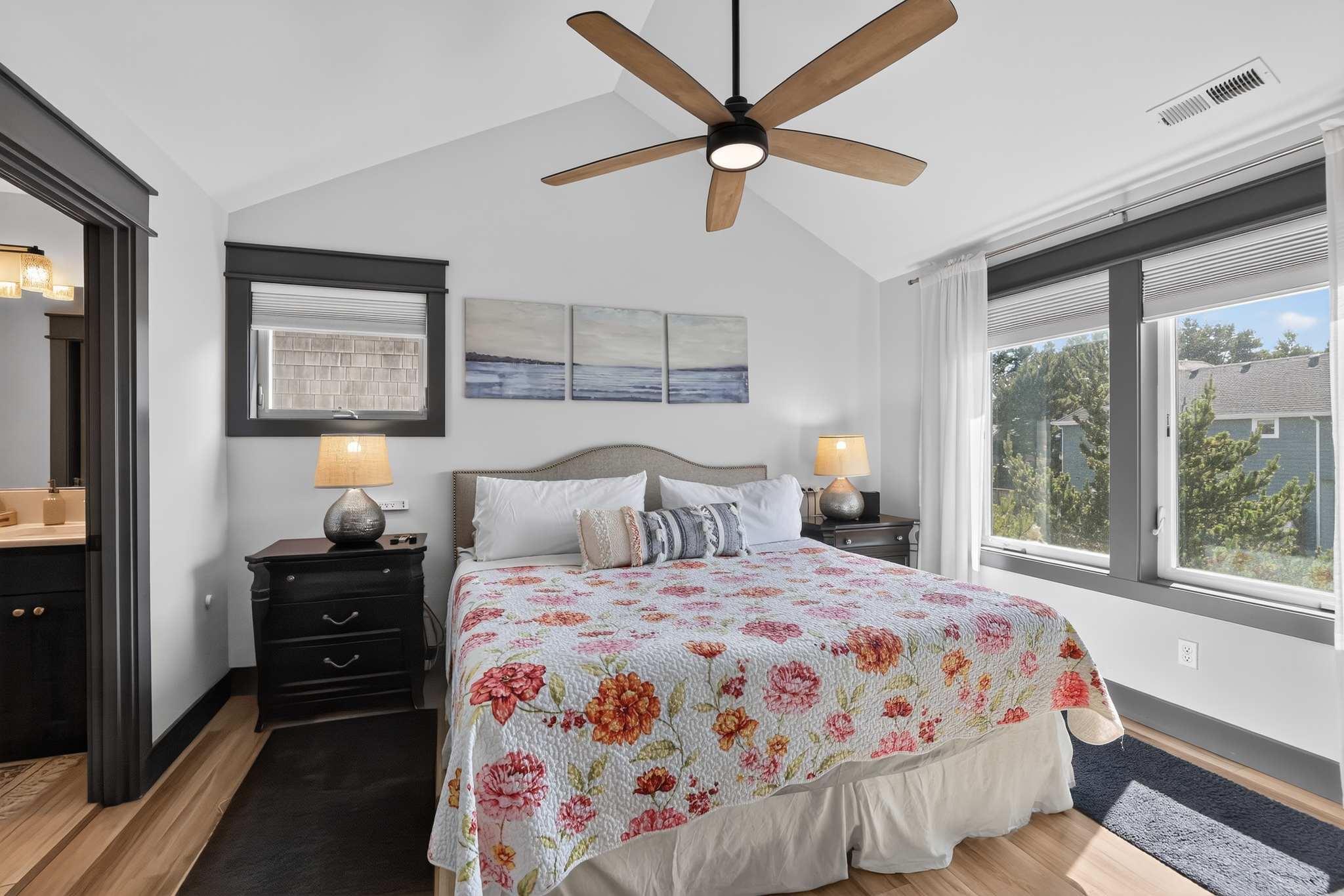 Cozy bedroom with a queen-sized bed adorned with a floral quilt, flanked by dark nightstands with lamps, and a large window offering views of the outdoors. A modern wooden ceiling fan hangs above.
