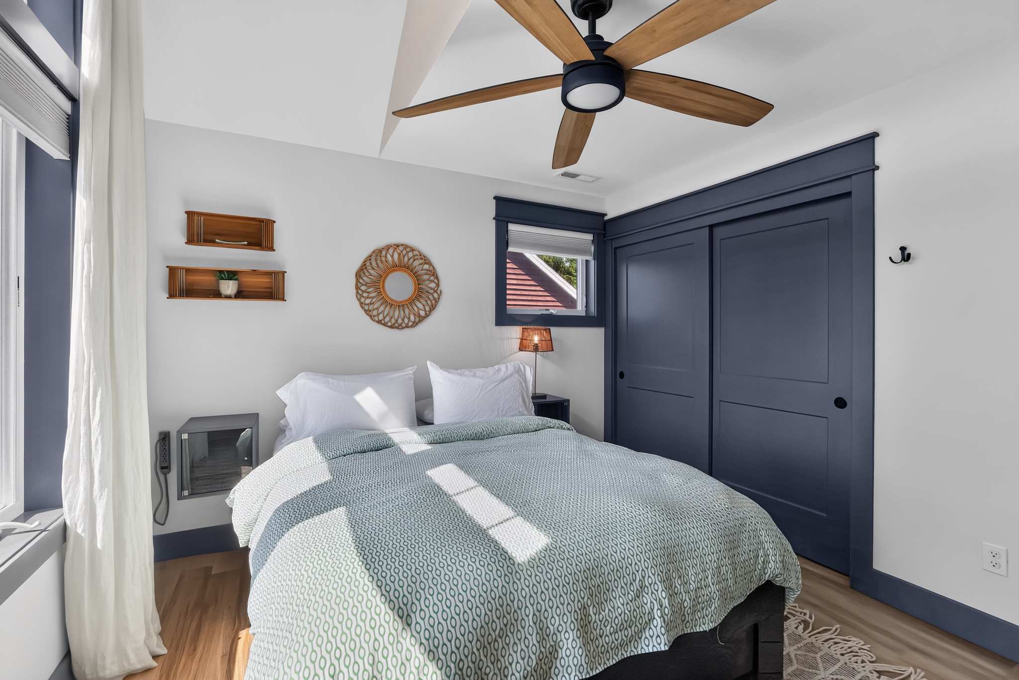 A bright bedroom features a queen-sized bed with a patterned green and white comforter, a wooden ceiling fan, and dark blue sliding closet doors, with natural light streaming in from a window.