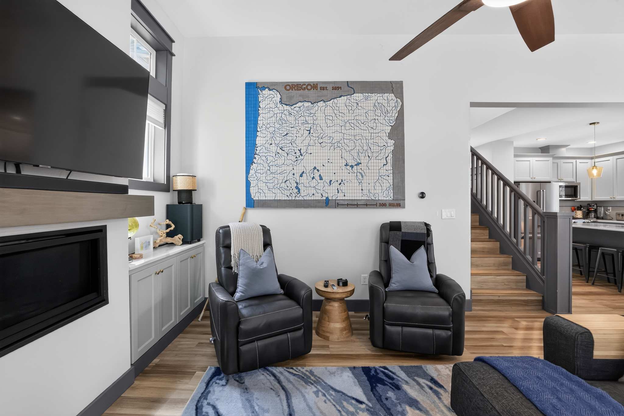 A cozy living room features two dark leather recliners facing a wall-mounted TV above a modern fireplace, with a large map of Oregon on the wall and a staircase visible in the background.