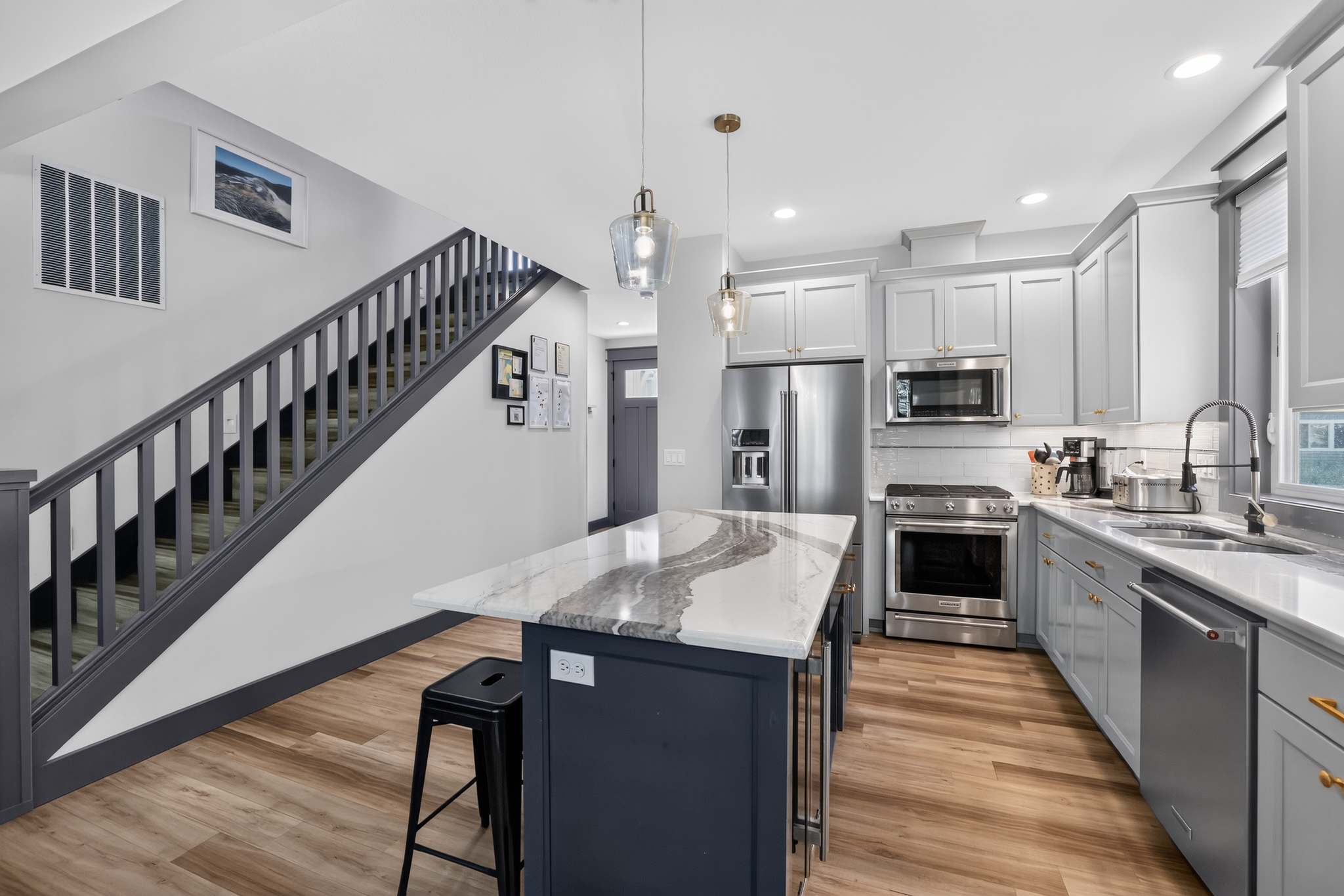 A modern kitchen features light gray shaker cabinets with gold hardware, stainless steel appliances, and a dark blue island with a white and gray marble-patterned countertop and bar stools. A staircase with dark gray railings is visible to the left.