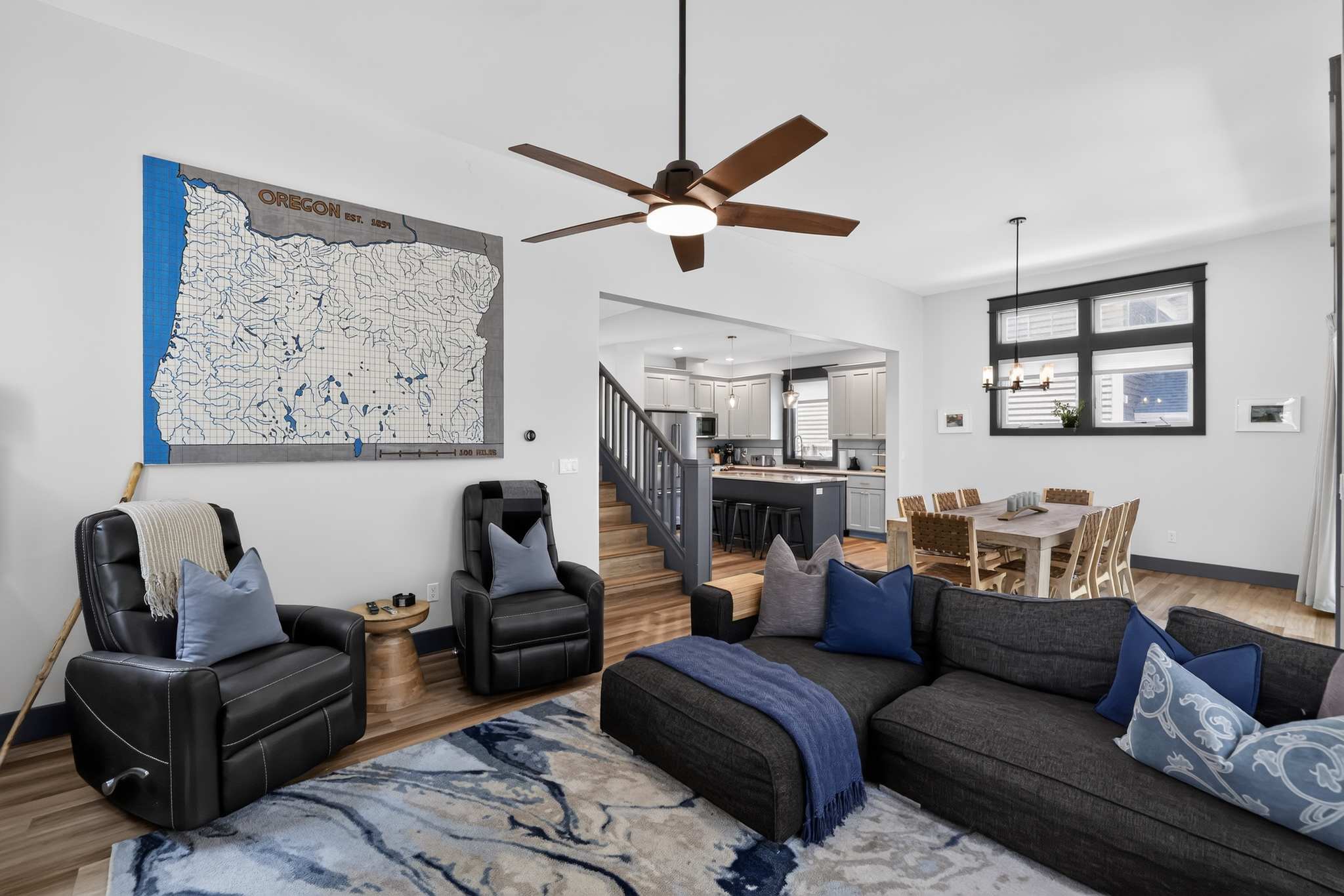 A bright open-concept living space features a dark gray sectional sofa with blue pillows, two black leather recliners, and a large Oregon map art piece on the wall, with a dining area and kitchen visible in the background. A ceiling fan with wooden blades hangs overhead.