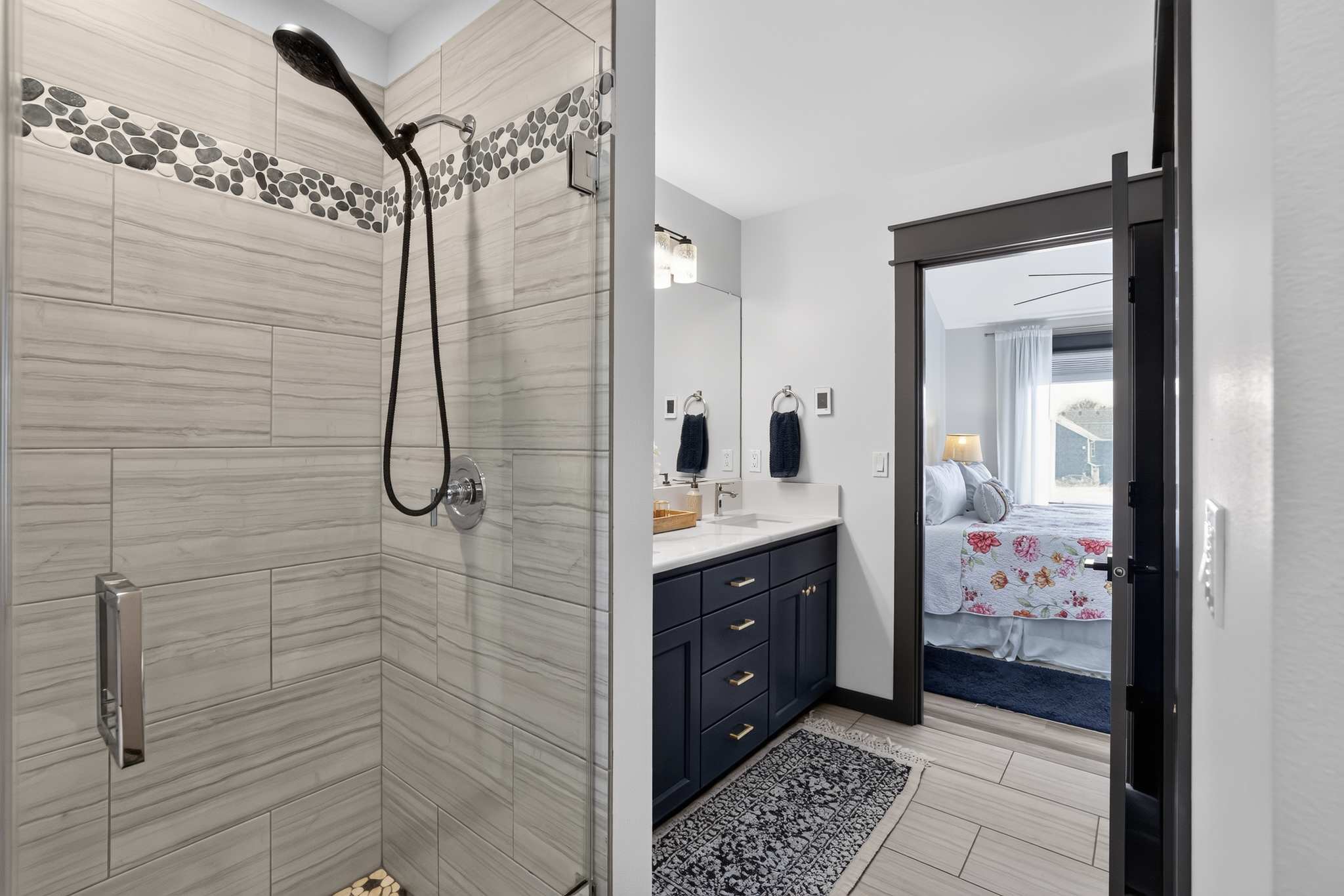Contemporary bathroom featuring a walk-in shower with light gray rectangular tiles and a decorative pebble mosaic accent strip, a dark blue vanity with a white countertop and gold hardware, and a view into an adjacent bedroom.