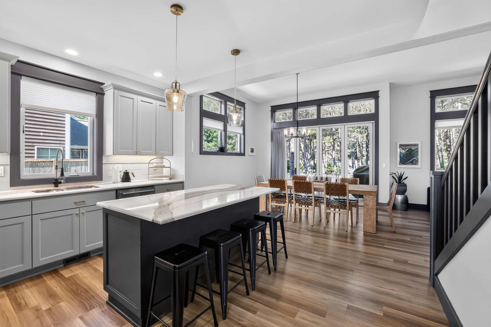 A modern open-concept kitchen features light gray cabinetry, white marble countertops, and a dark gray island with black bar stools, illuminated by brass pendant lights. Adjacent is a dining area with a long wooden table and leather chairs, set against large windows overlooking trees.