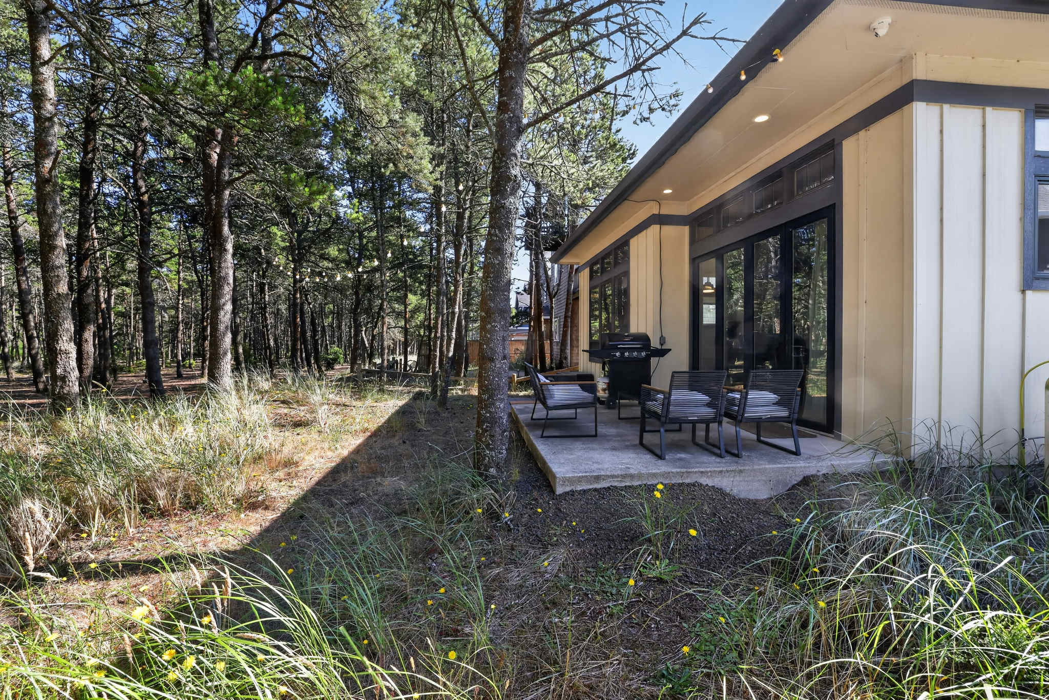 A concrete patio extends from the light yellow house, furnished with a gas grill and a set of four outdoor chairs around a small table, all set against a backdrop of dense pine trees and natural ground cover.