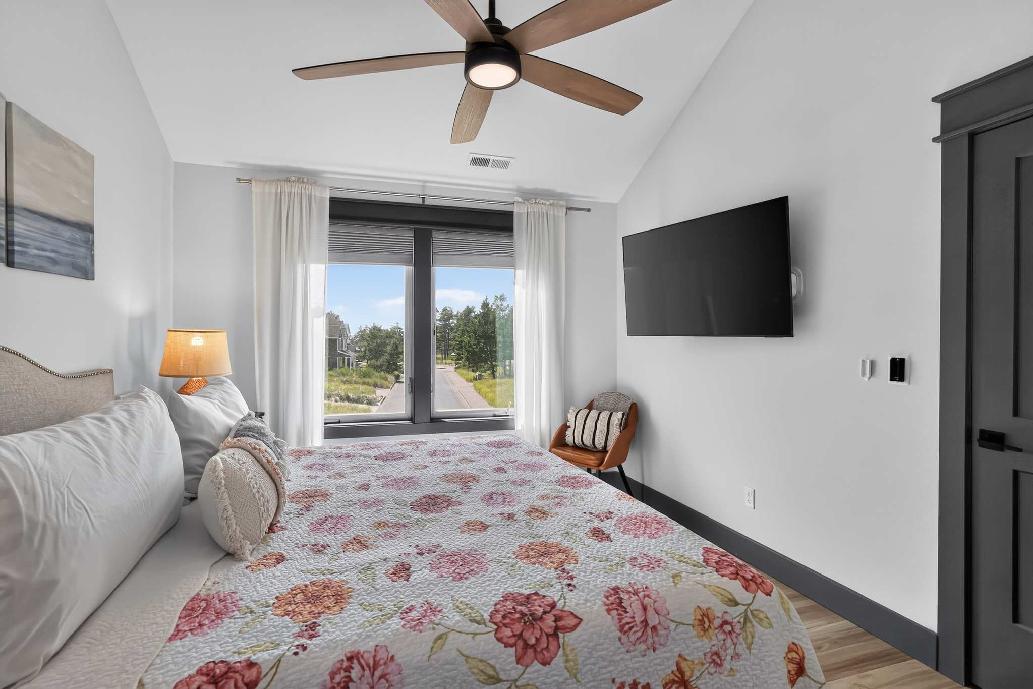 View of bedroom-1 showing the queen-sized bed with a floral quilt, a wall-mounted television, and a large window with white blinds and curtains offering an outdoor view. A modern wooden ceiling fan is visible overhead.