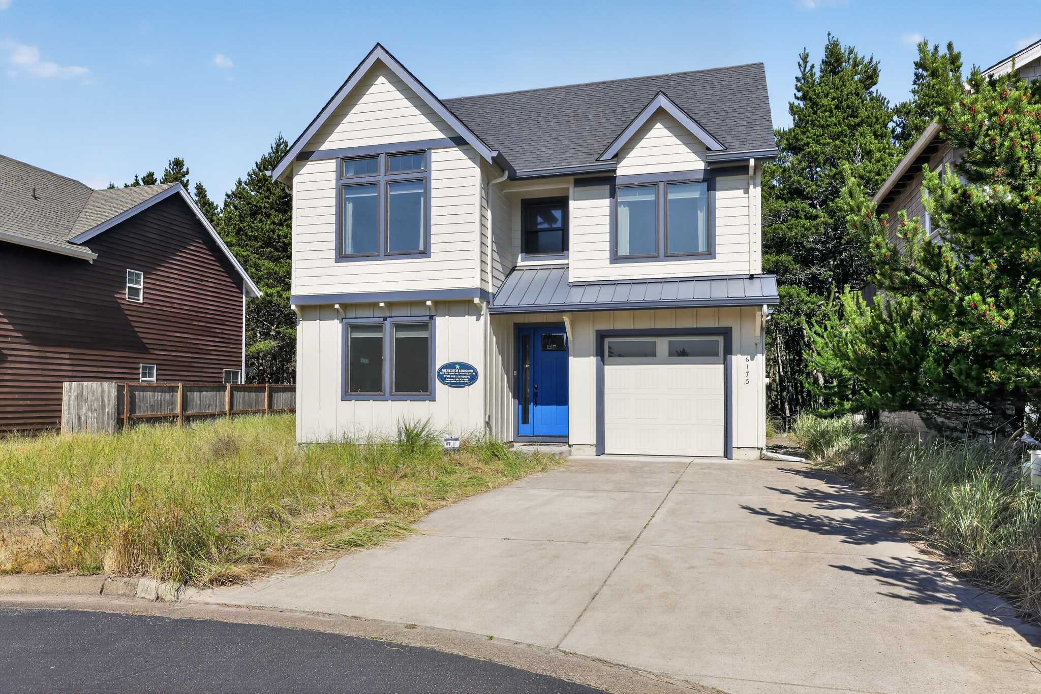 A charming two-story house with light-colored siding and dark blue trim features a bright blue front door, a single-car garage, and a concrete driveway, set against a backdrop of green trees and a clear blue sky.