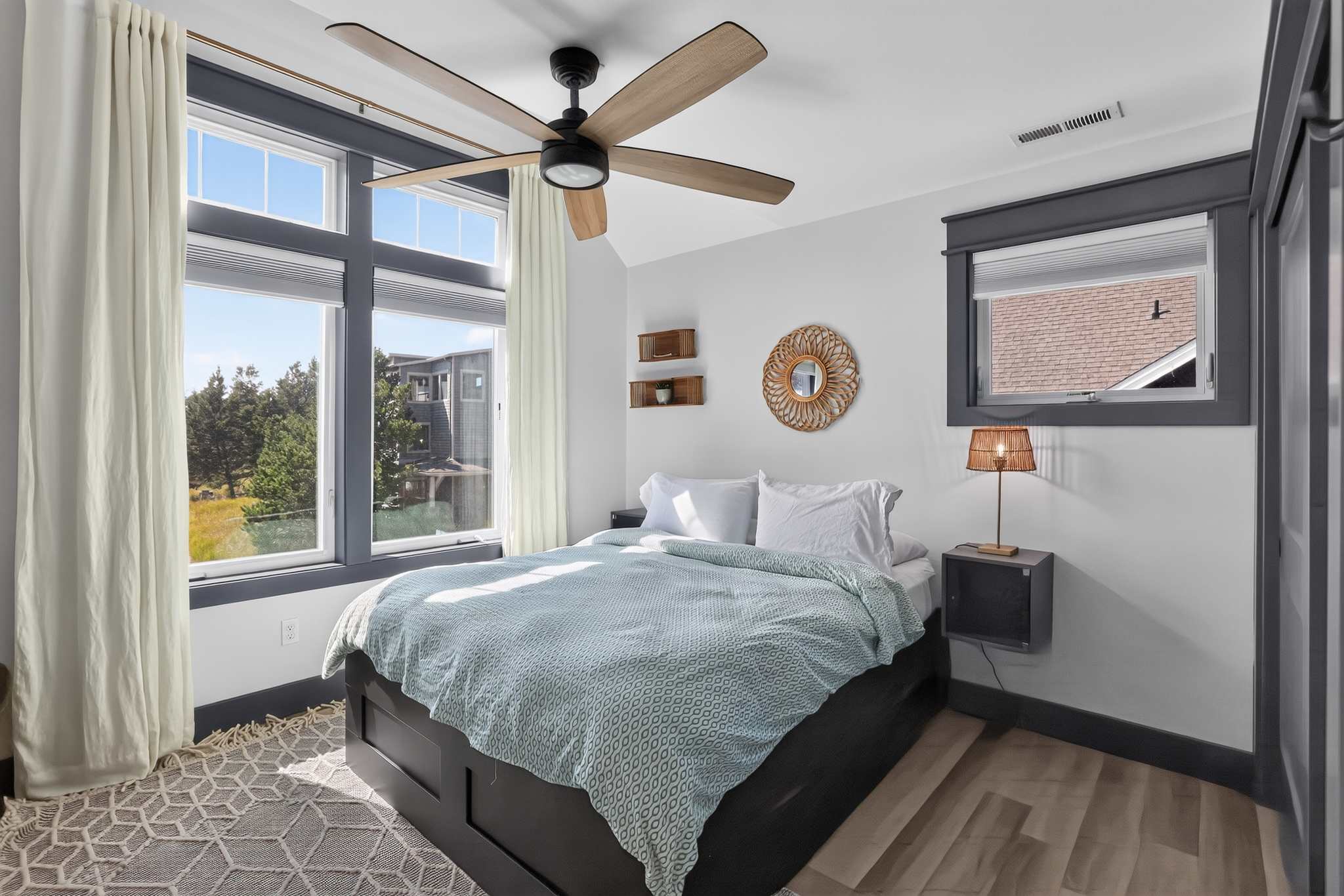 Bright bedroom with a queen-sized bed featuring a light blue patterned duvet, a large window with white curtains, and a modern wooden ceiling fan. A small round mirror and floating shelves decorate the wall.