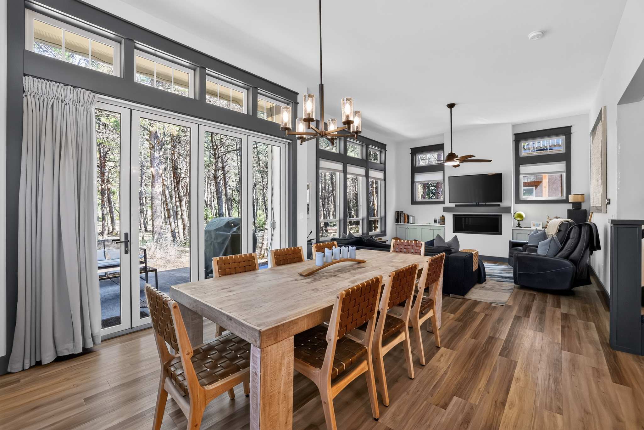 Bright dining area with a large rustic wooden dining table and eight woven leather chairs, situated next to expansive glass doors that open to an outdoor patio. The room connects to the living room, visible in the background, and features large windows with dark trim.