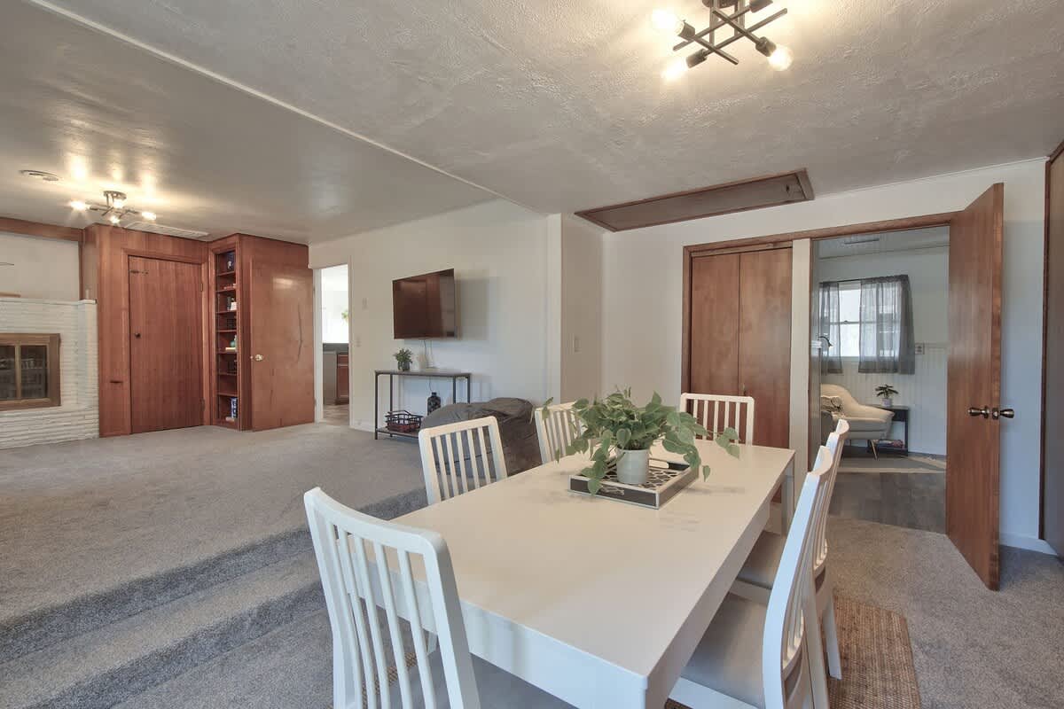 Bright dining area with a white table and chairs, featuring a modern light fixture, and stepping down into a carpeted living space with a wall-mounted TV and a glimpse of a white brick fireplace.