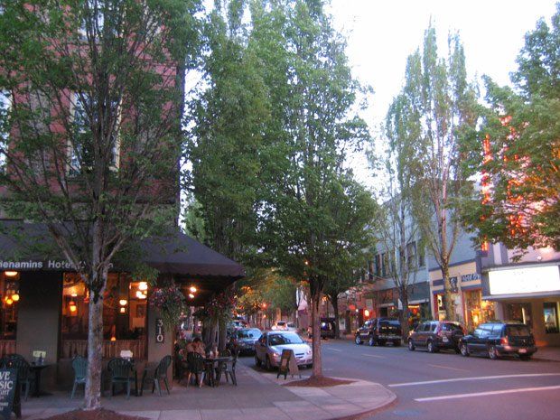 An evening street scene in a charming town features cars parked along the curb, buildings with illuminated windows and awnings, and several groups of people enjoying outdoor dining. Mature trees line the sidewalk, casting soft shadows.