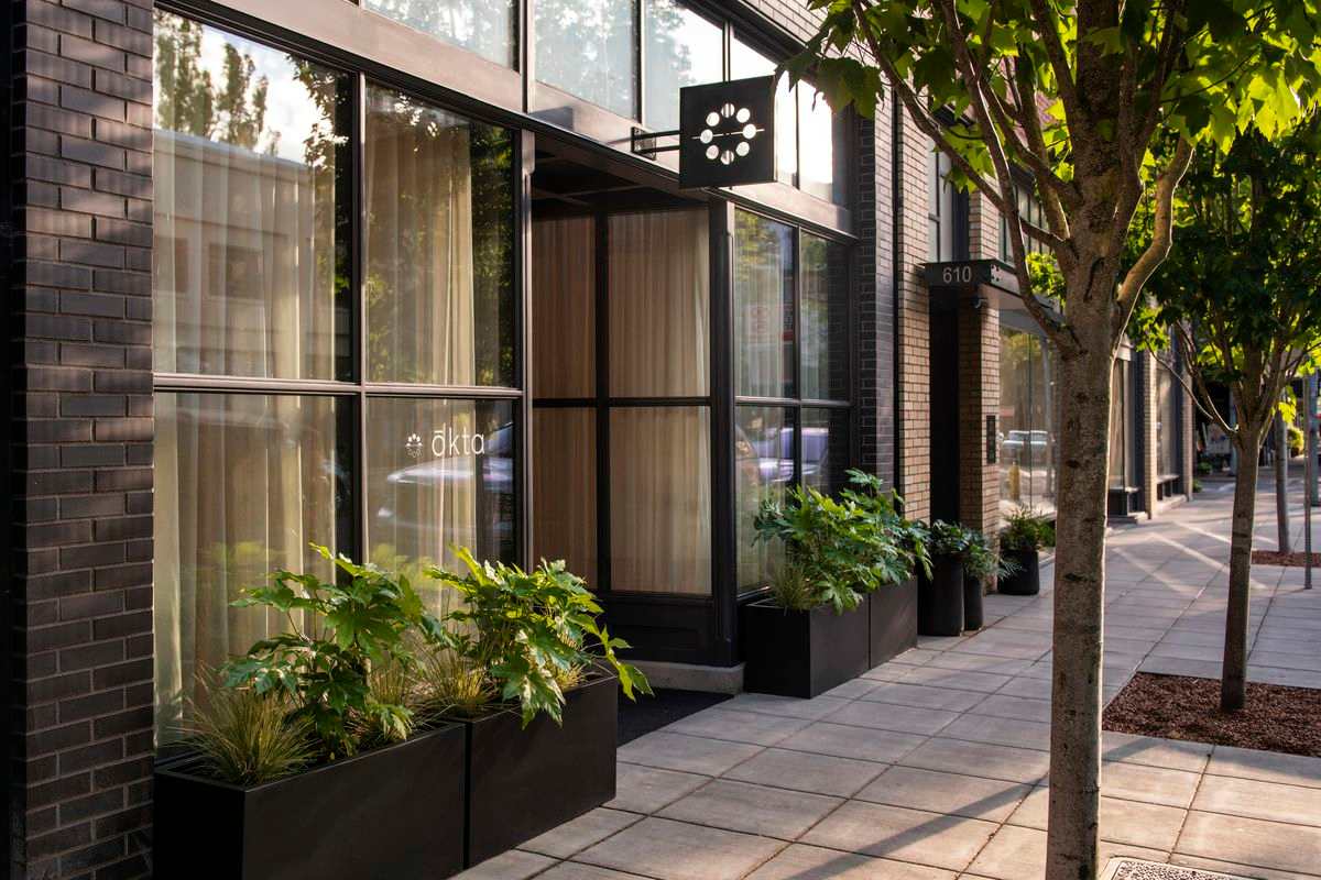 A sleek, modern building entrance features large glass windows, black brick facade, and a row of dark planters filled with lush green foliage along the sidewalk. A small, geometric sign hangs above the main doorway.