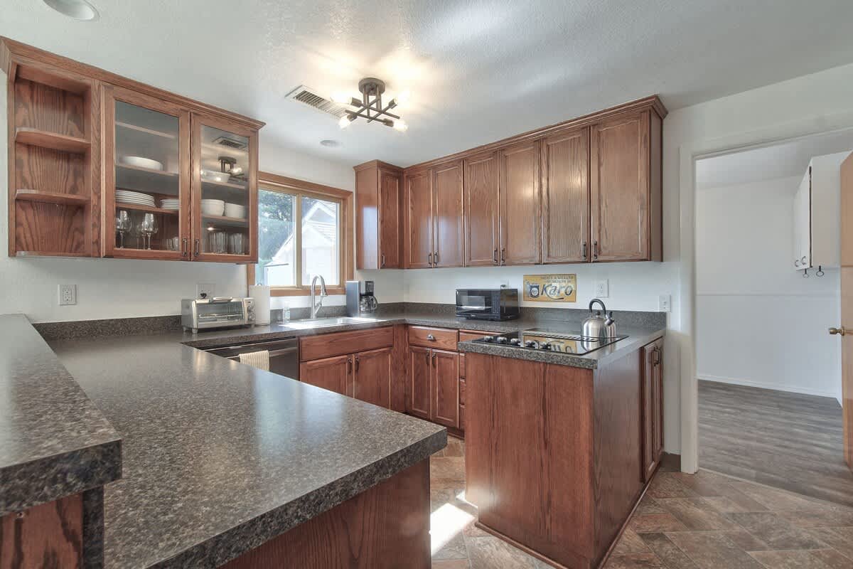 A well-lit kitchen with dark wood cabinets and dark speckled countertops, featuring a stainless steel dishwasher and a built-in electric cooktop. A window above the sink provides natural light, and glass-front cabinets display dishware.