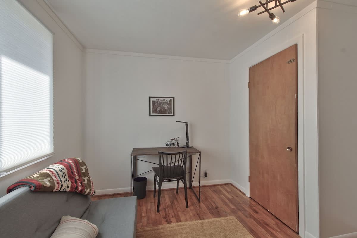 Compact and functional office space featuring warm hardwood floors, a simple metal-framed desk with a black chair, a grey sofa/daybed, and a window with a cellular shade.